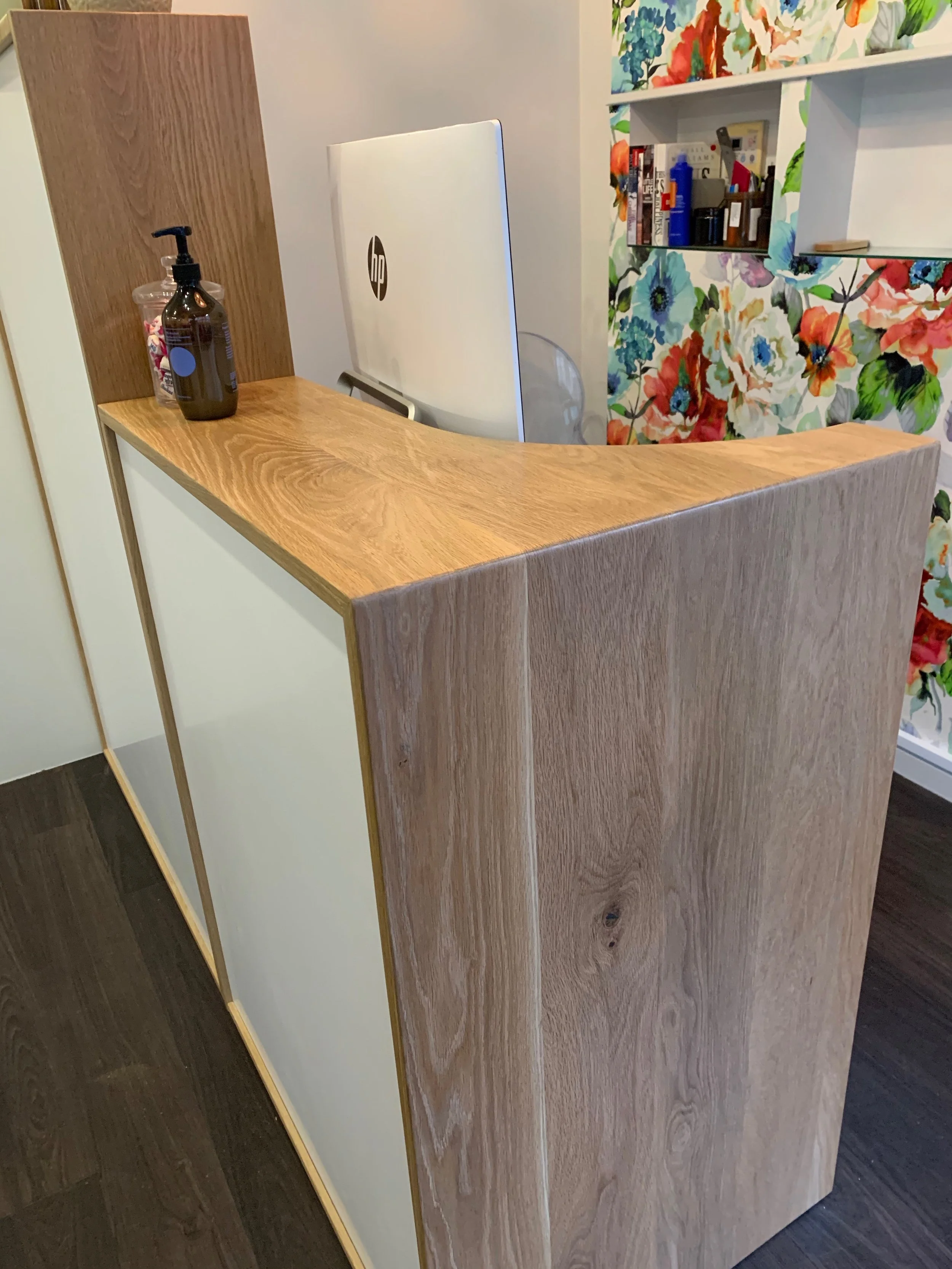 White melteca and oak reception desk. HP computer visible on desk as well as product bottle and jar of lollies