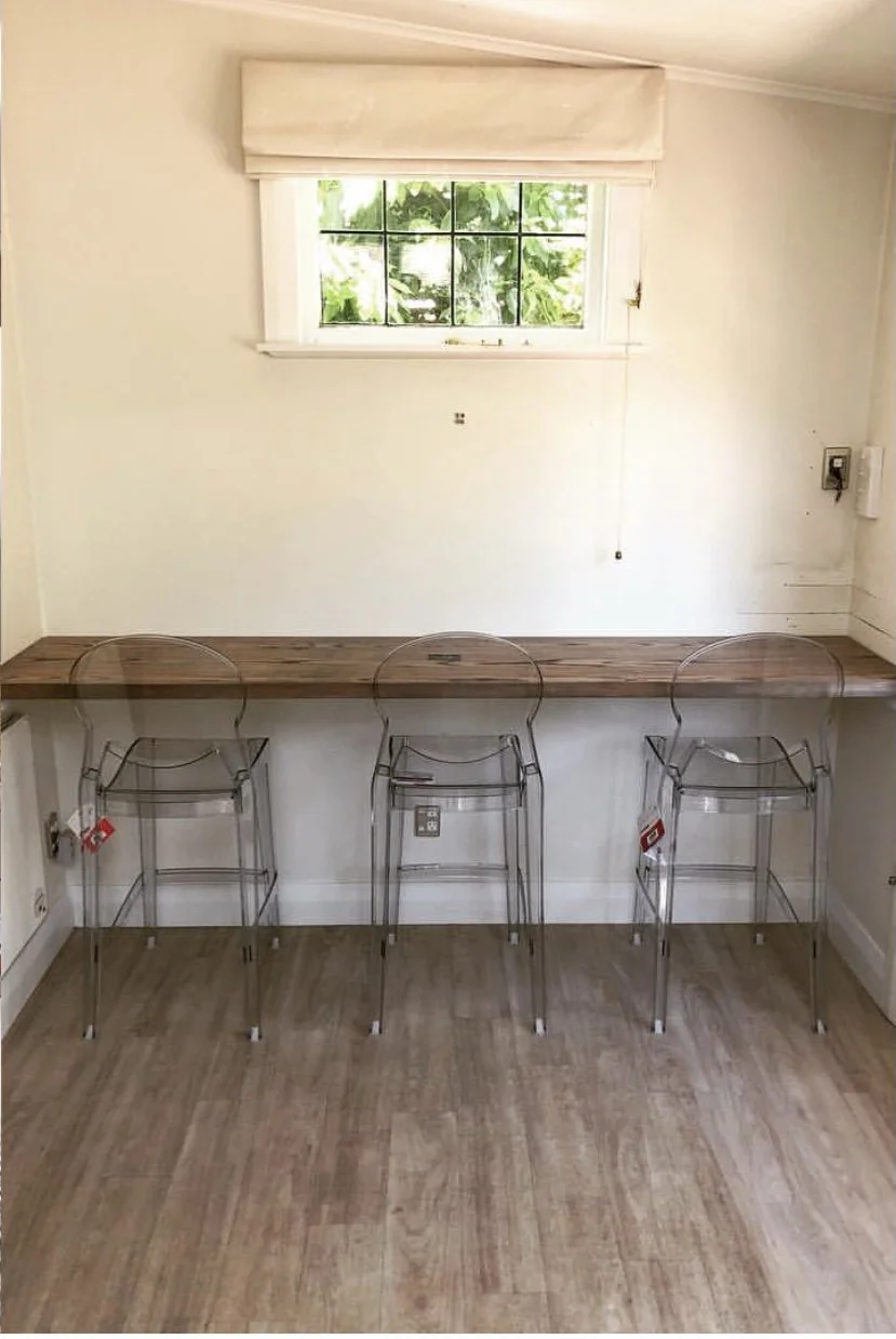 Heavily ghost washed American ash kitchen breakfast bar with three clear plastic chairs seated at benchtop. a window above has a beige roman curtain 