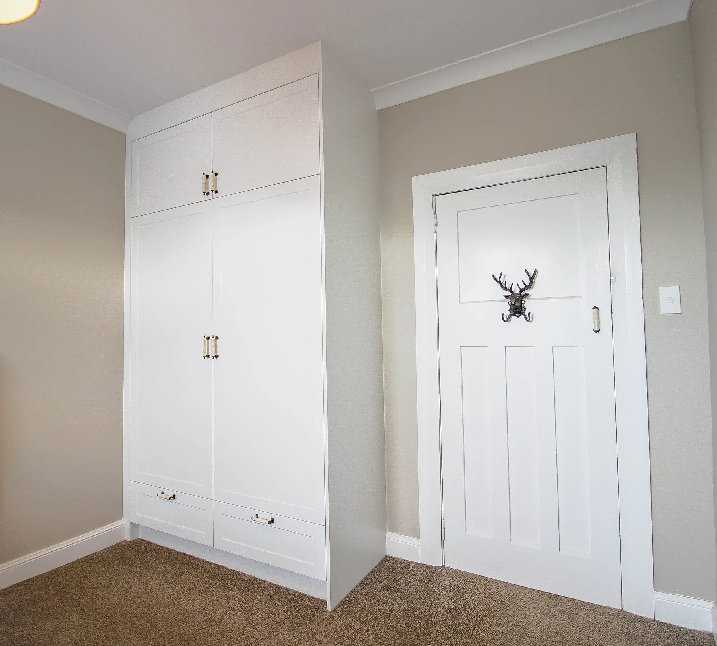 White built in panel wardrobe with handles in a dark beige room. A white door with a deer shaped coat hook hangs on the back of the closed door