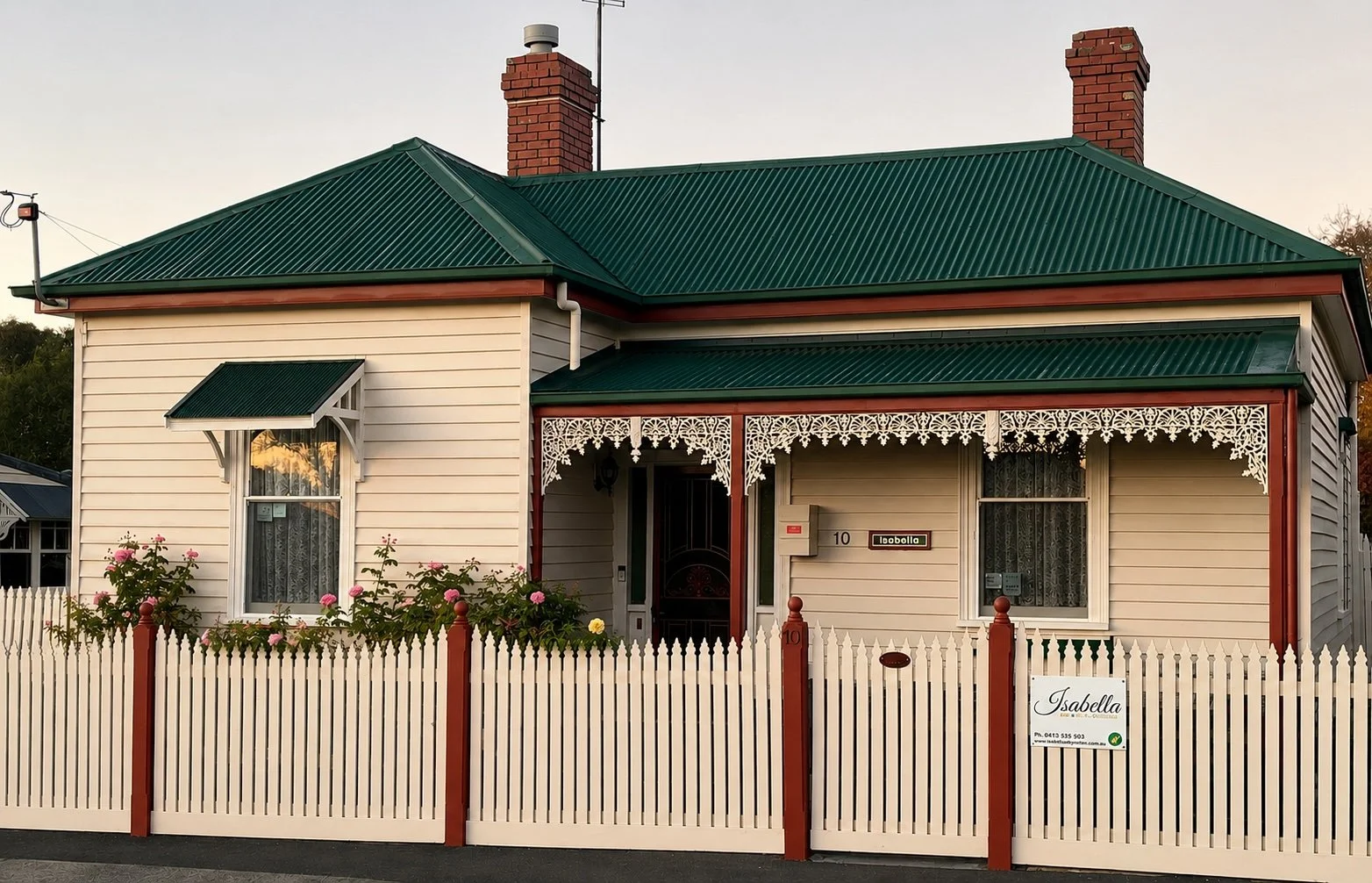 A house with cream-colored siding, a green metal roof, and a white picket fence. There is a decorative lace-like trim on the porch, with a sign that reads 'Isabella' and a phone number.