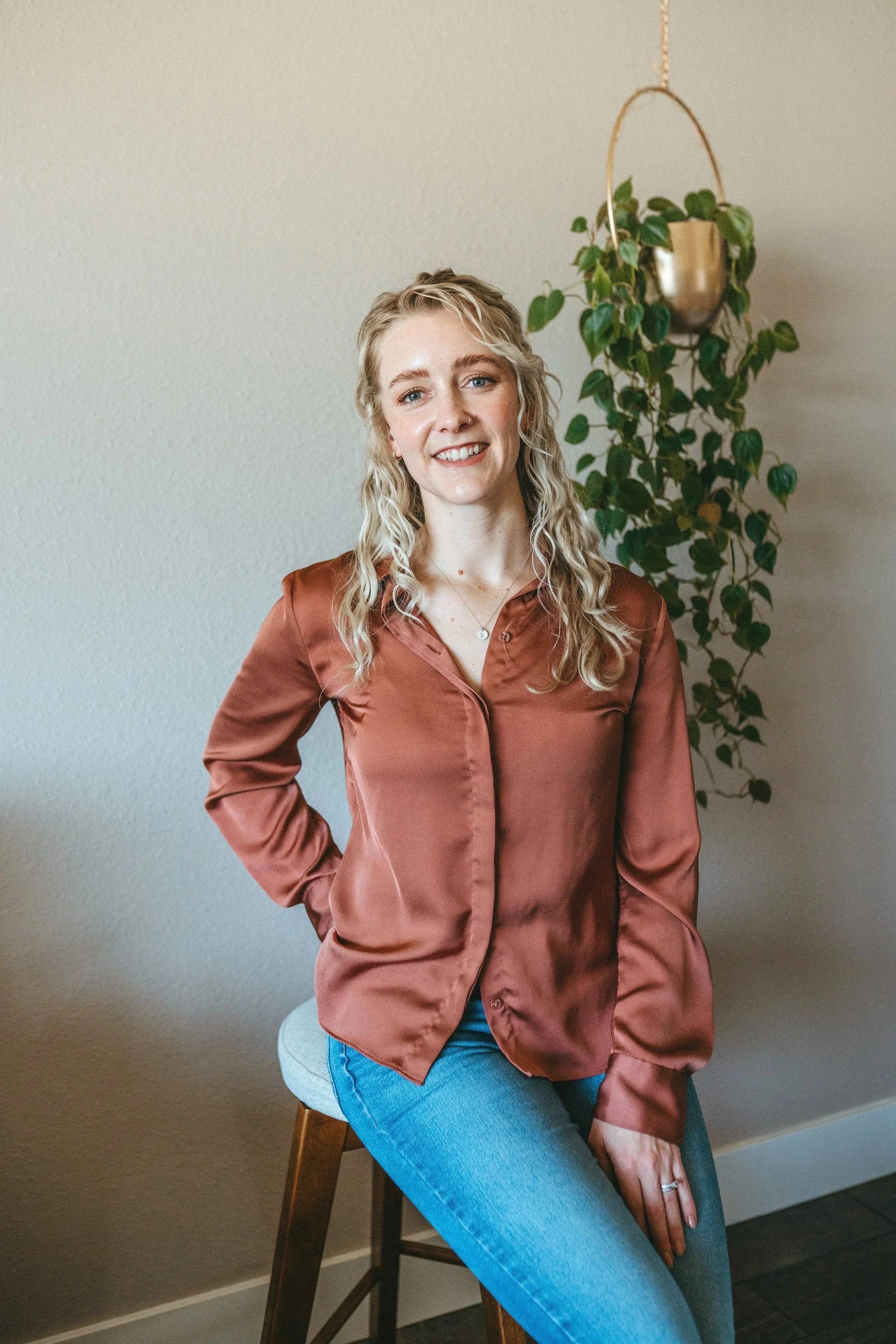 A woman with wavy blonde hair and fair skin, wearing a rust-colored satin blouse and blue jeans, sitting on a wooden stool with a light gray cushion. Behind her is a hanging potted plant on a metallic stand against a beige wall.