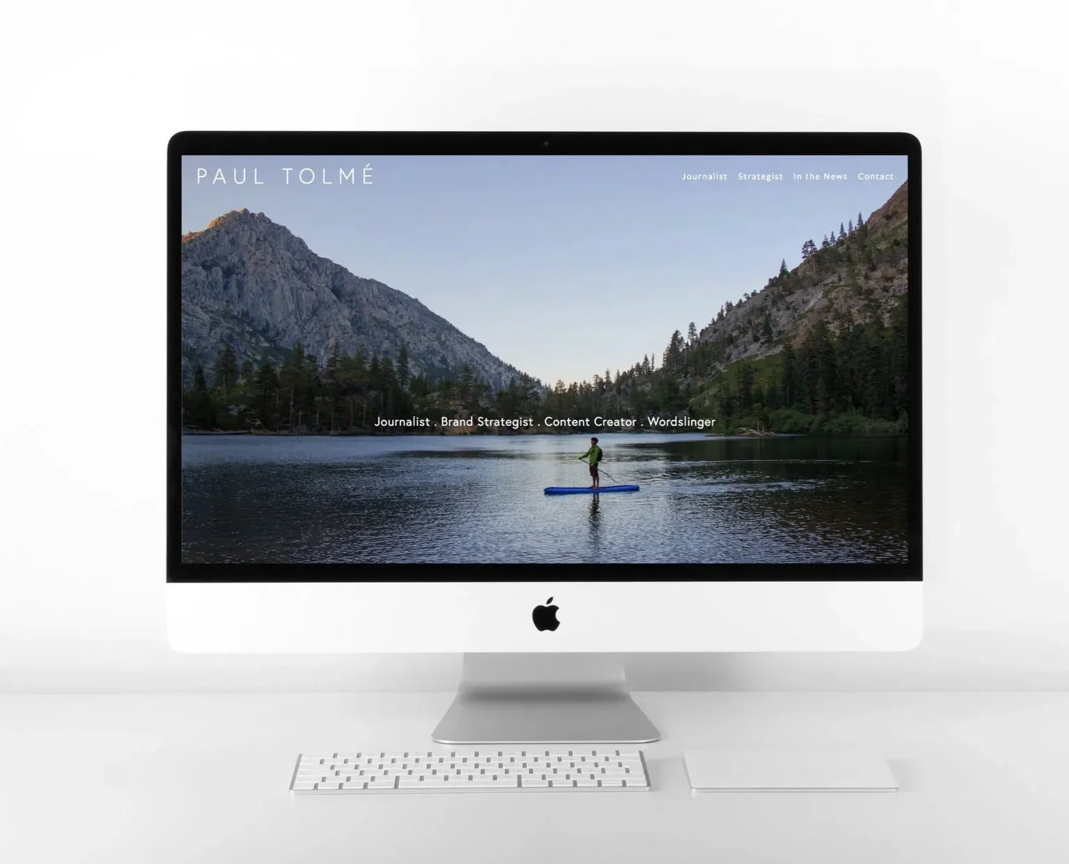 Apple iMac computer on white desk displaying a landscape photo of mountains and a lake with a person on a paddleboard.