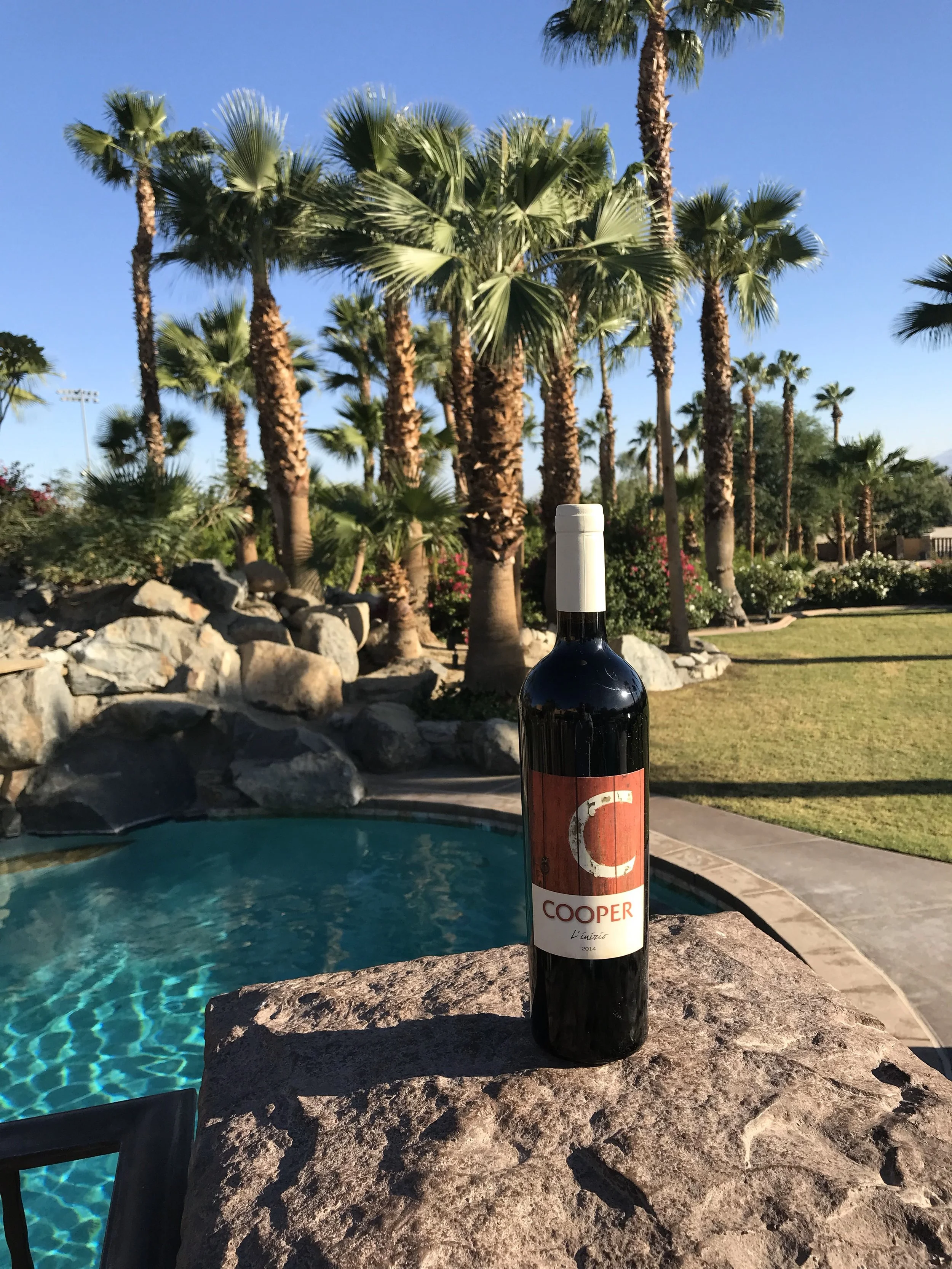 Bottle of red wine labeled 'Cooper,' placed on a stone surface outdoors next to a pool with palm trees in Palm Springs CoopRV and a lawn in the background.