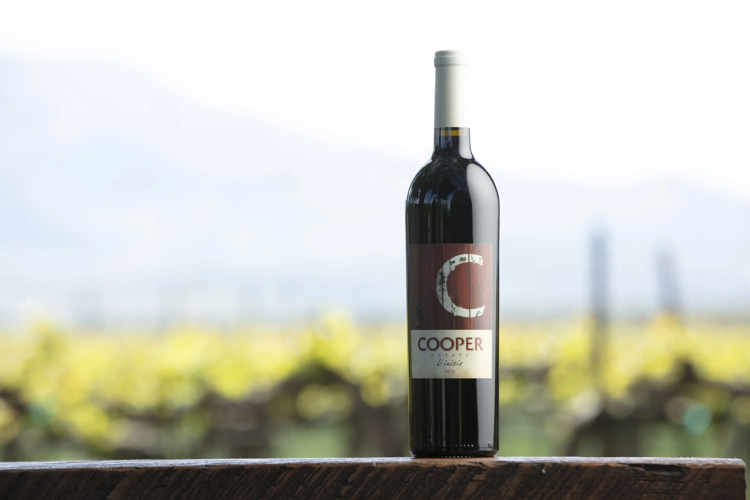 A bottle of red wine at Red Mountain, Washington with a red and white label reading 'Cooper' and 'Estate,' standing on a wooden surface with a vineyard in the blurred background.