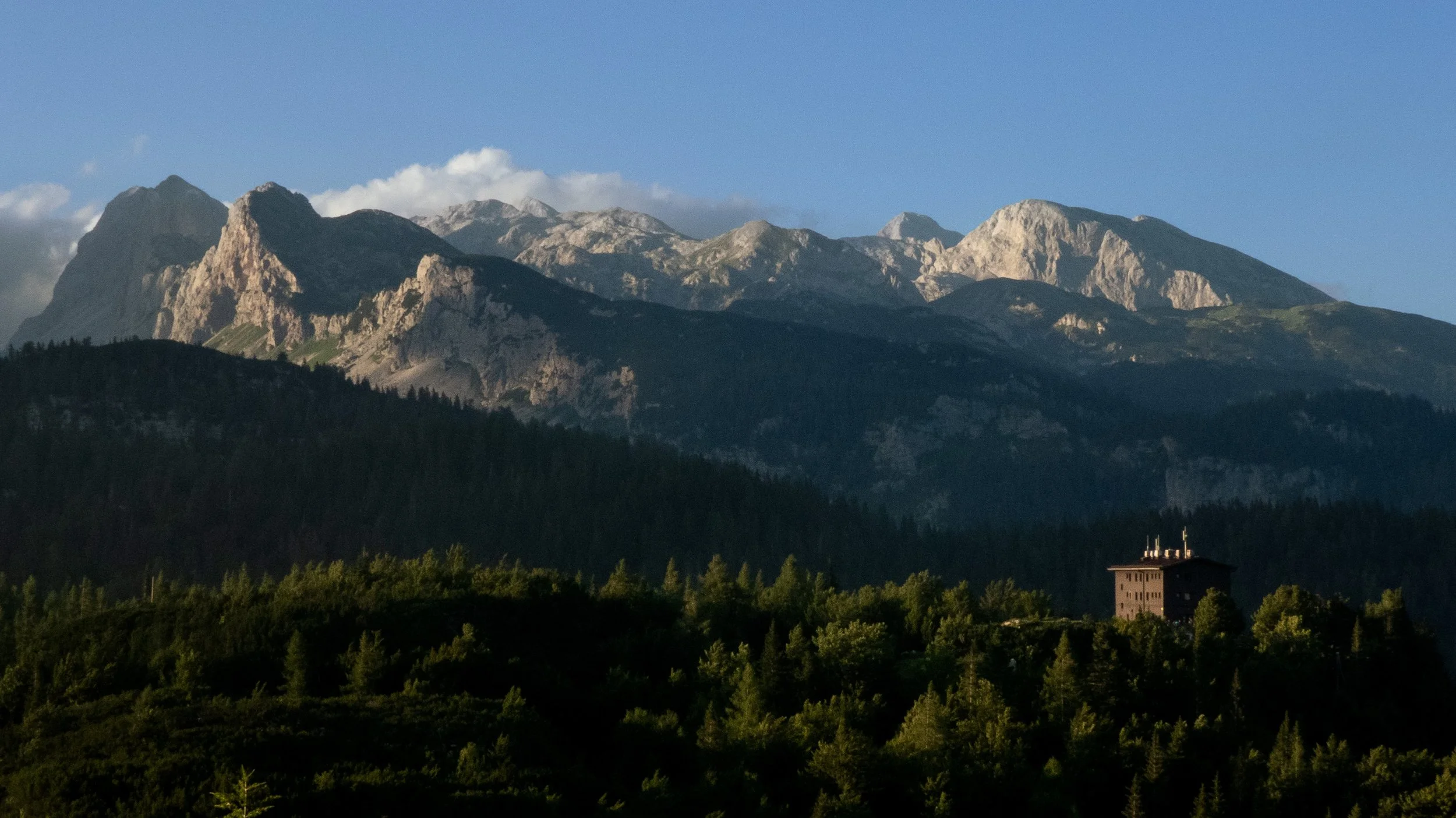 Mountain range with rocky peaks, forested hills in the foreground, and a building on the hill.
