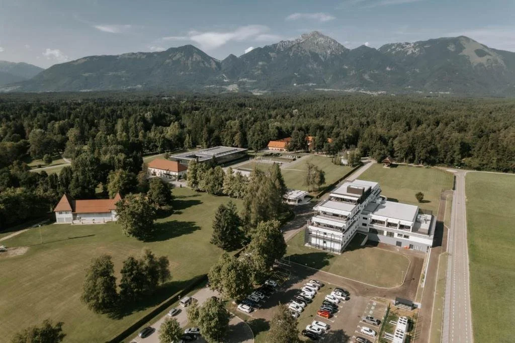 An aerial view of a large complex with modern white buildings, surrounded by green lawns, trees, and parking lots, with a mountain range in the background under a partly cloudy sky.