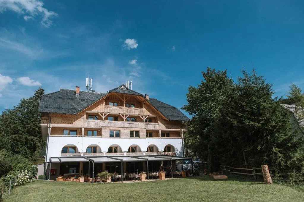 A multi-story building with wooden and white exterior walls, large balconies, and a dark roof, set against a bright blue sky with some clouds, surrounded by green trees and a grassy area with outdoor seating.