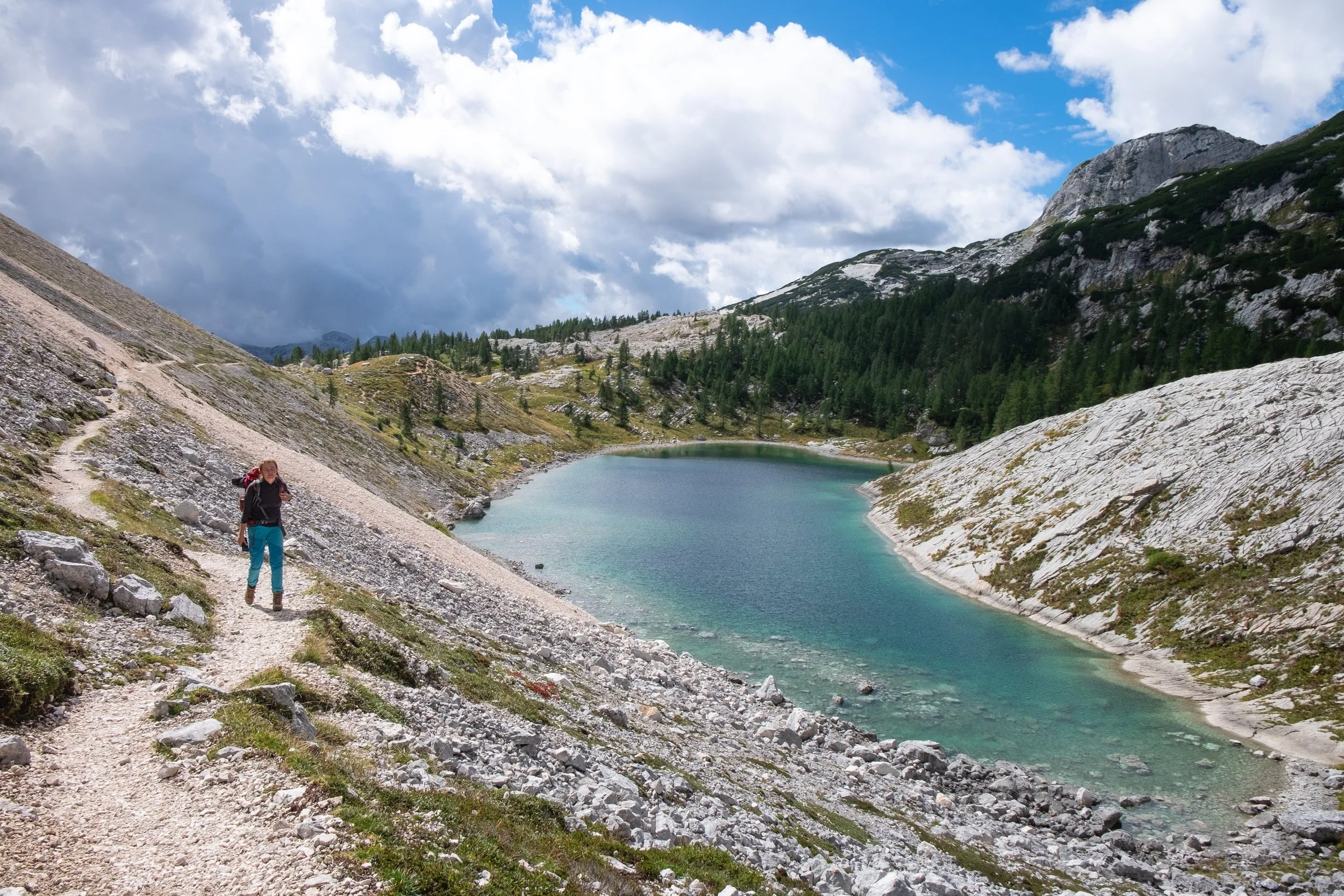A person hiking on a trail beside a mountain lake in a mountain landscape with rocky slopes, green trees, and a cloudy sky.