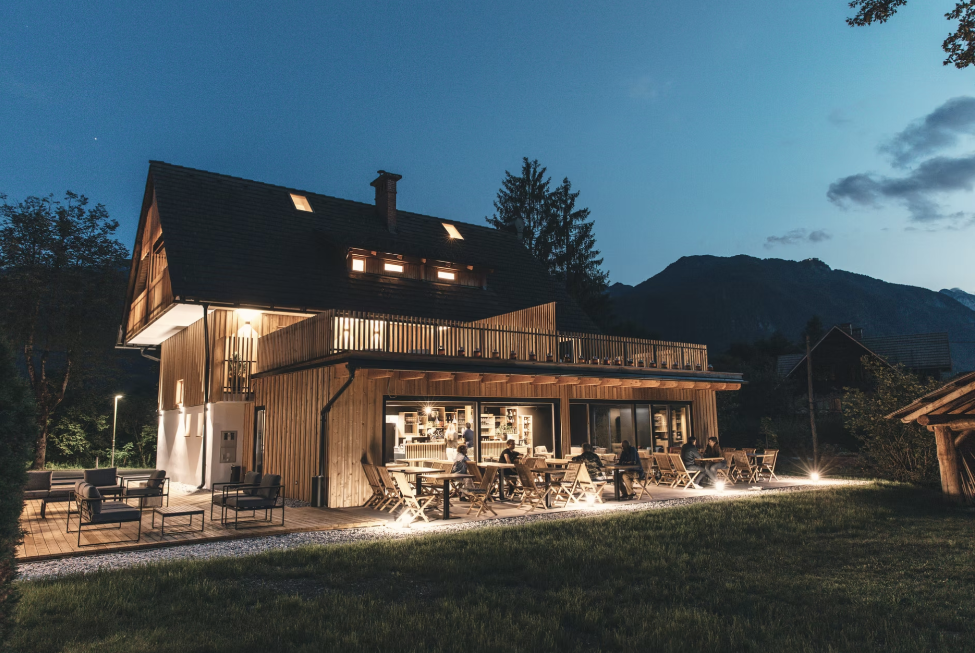 A wooden house with outdoor seating on a deck during nighttime, with lights illuminating the area, surrounded by trees and mountains in the background.