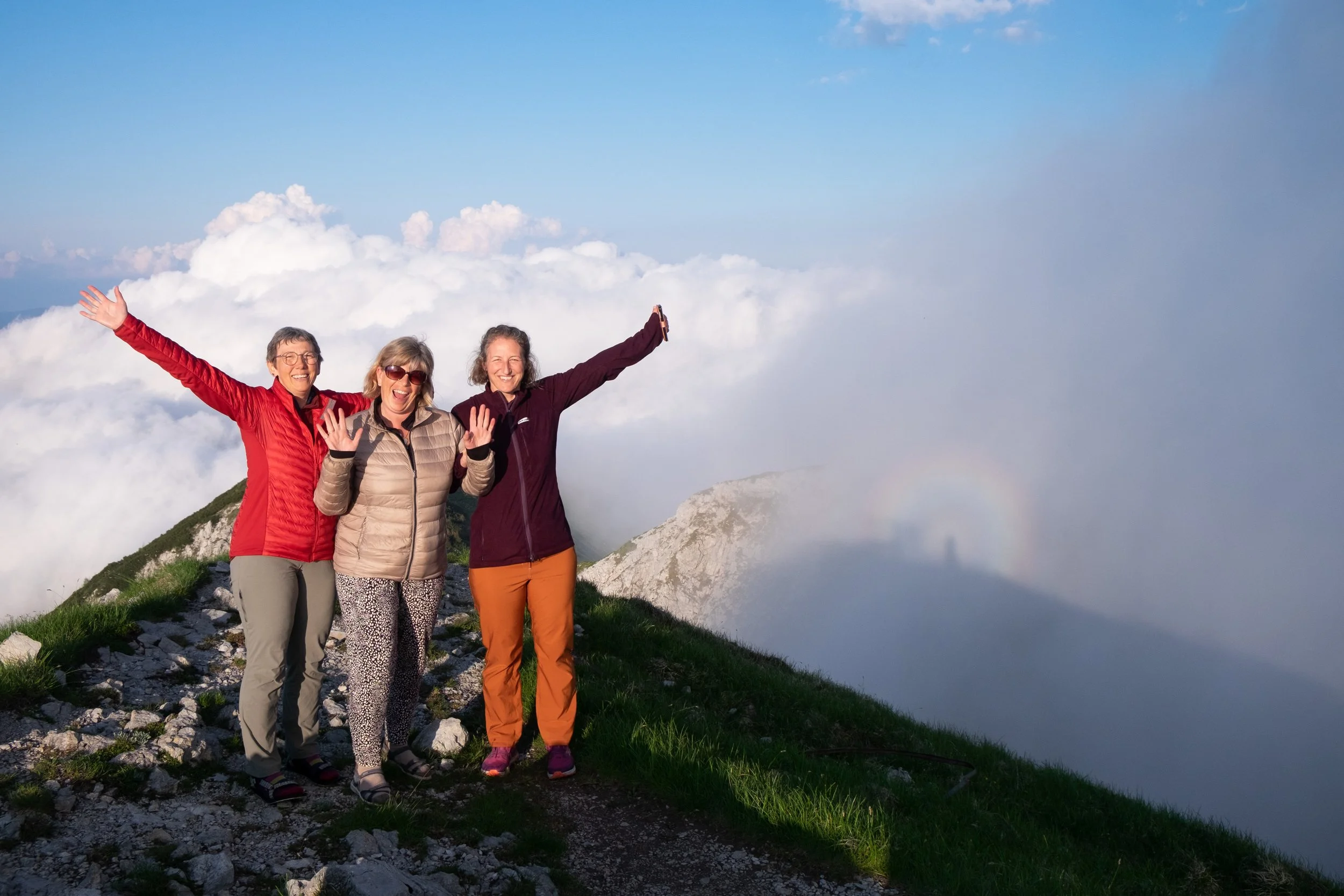 Three women standing on a mountain peak among clouds, smiling and raising their arms in celebration, with a rainbow faintly visible in the background.