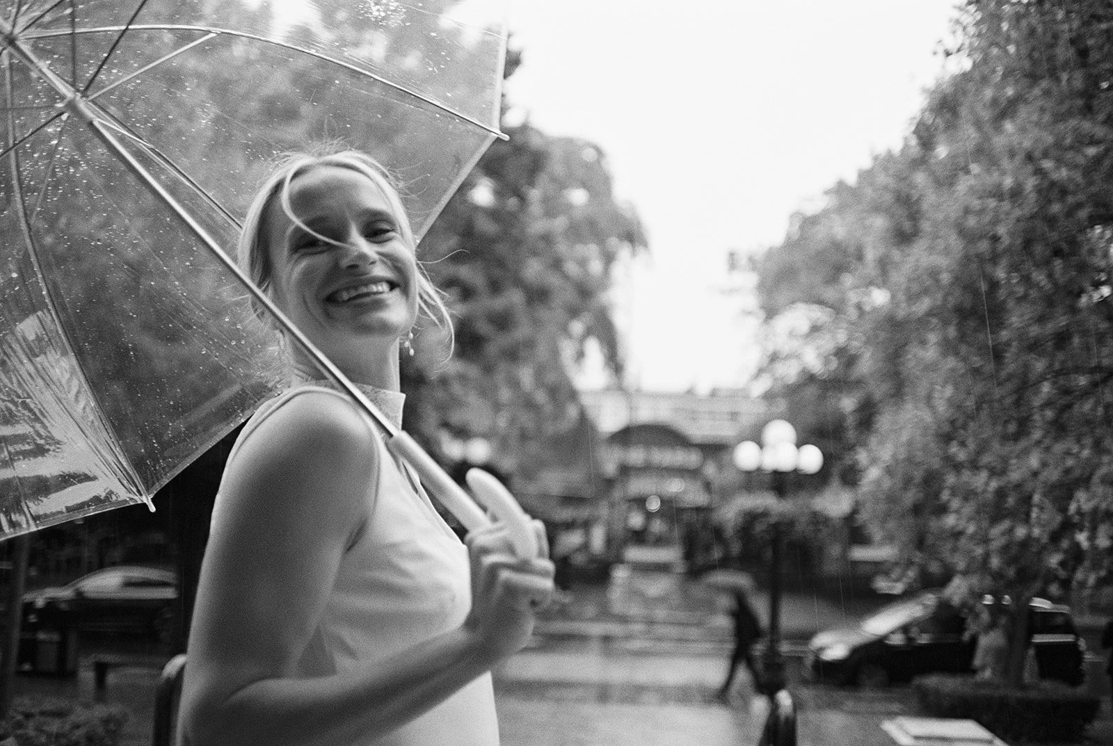 Smiling bride with a clear umbrella on a rainy day, captured by a Paris film wedding photographer amid trees and city buildings.