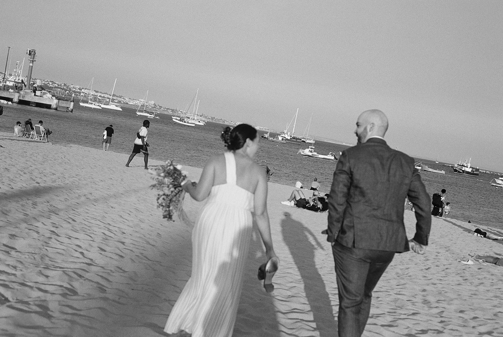 Bride and groom walk on Lisbon beach towards sea captured in black and white on film.