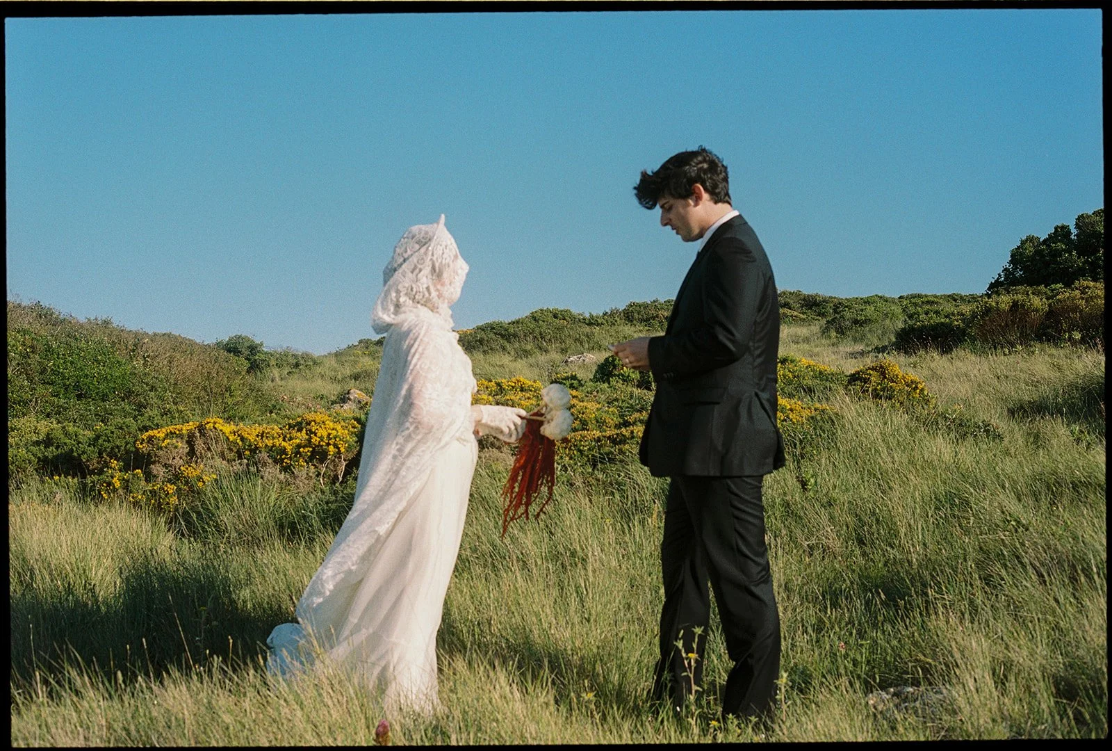 Warm, nostalgic film shot of bride in a white dress faces her groom in a grassy field and blue skies.