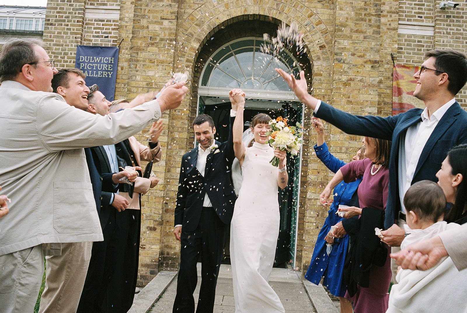 Guests throw flowers while bride and and groom walk outdoors smiling.