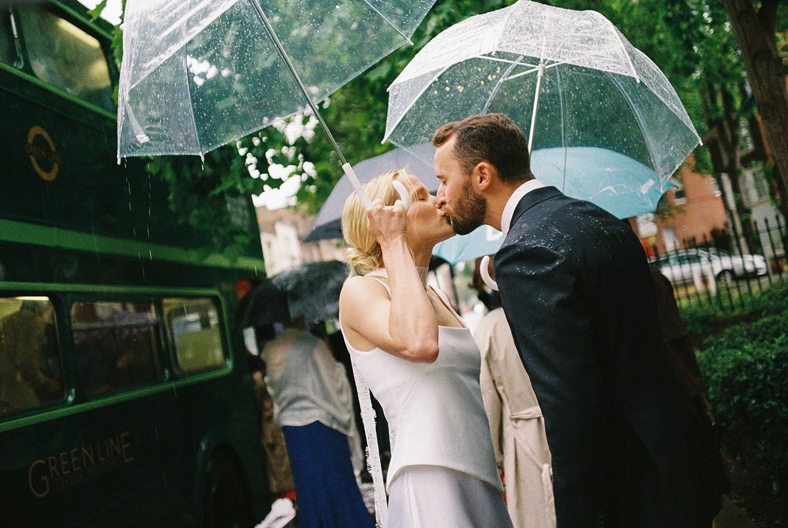 Bride and groom under clear umbrellas on rainy day in front on green bus.