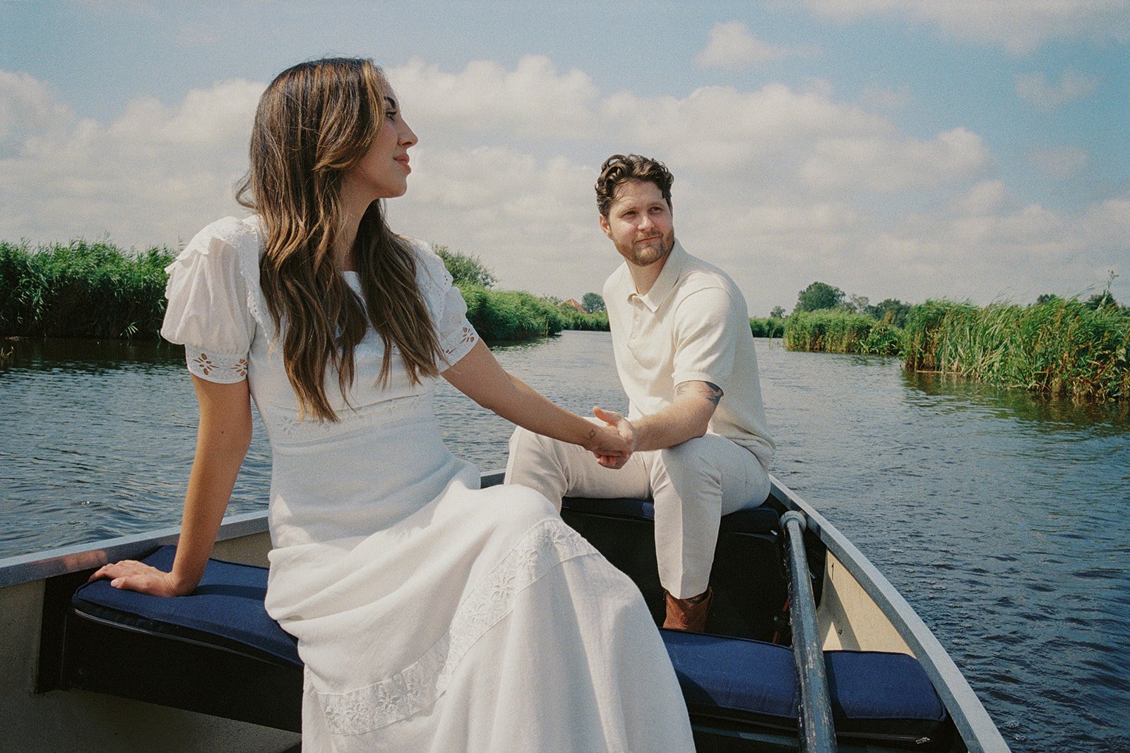 A bride and groom sit in a small boat on a calm river, holding hands on a sunny day.