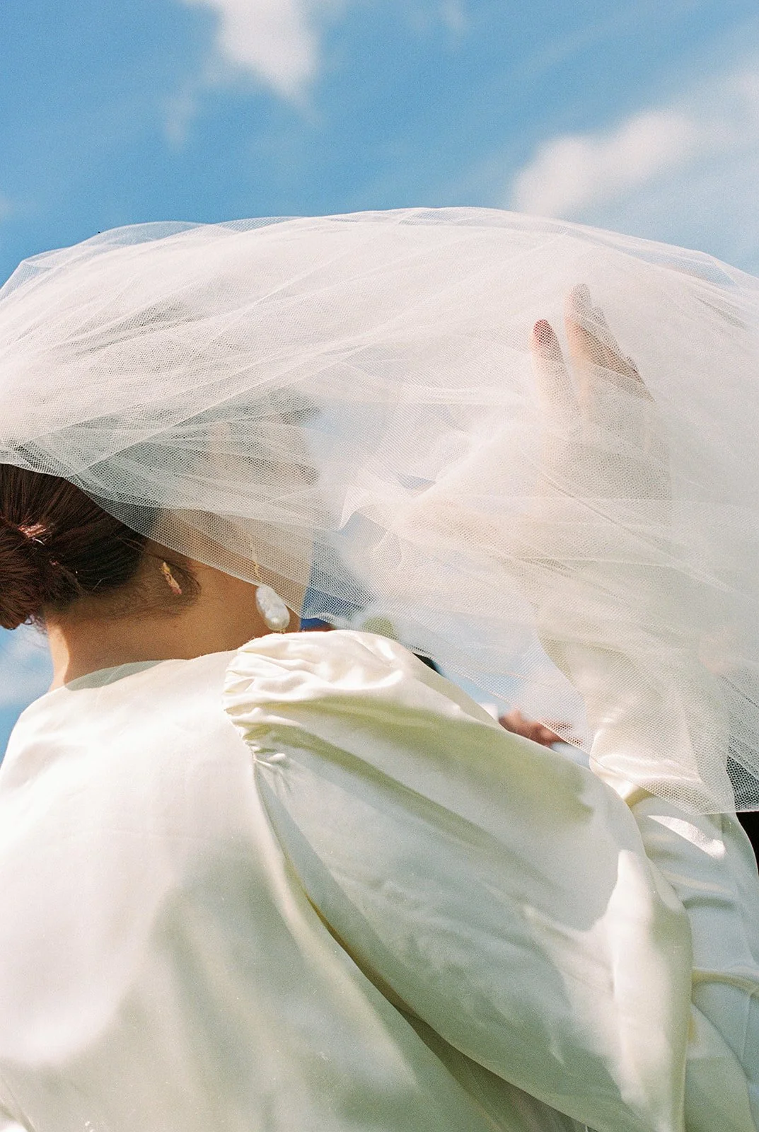 A bride in a white dress holds her veil under blue skies.