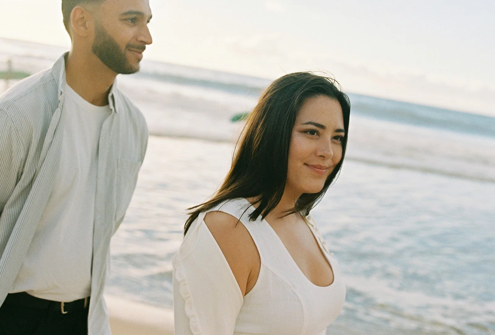 Bride smiling and groom walk together by the beach, with ocean and sky behind.