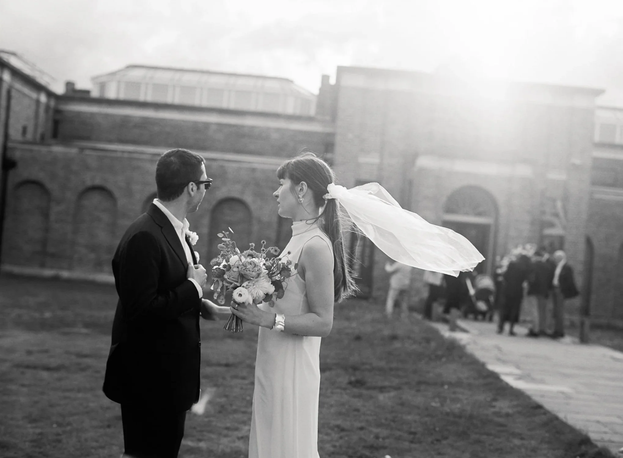 Artistic portrait of bride veil flowing on air while groom looks at wedding guests by London Film Wedding Photographer