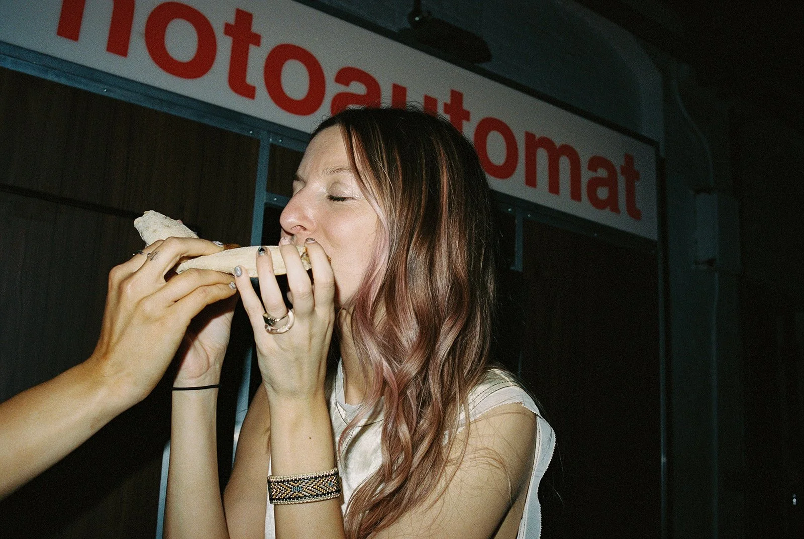 Bride is about to bite into a pizza slice, held by another guest, near a photoautomat booth.