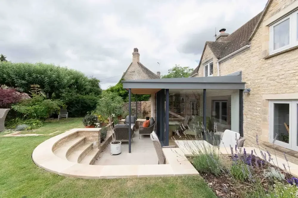 Contemporary glass and zinc garden room extension on a historic stone house, featuring a curved stone patio wall and integrated outdoor living space.