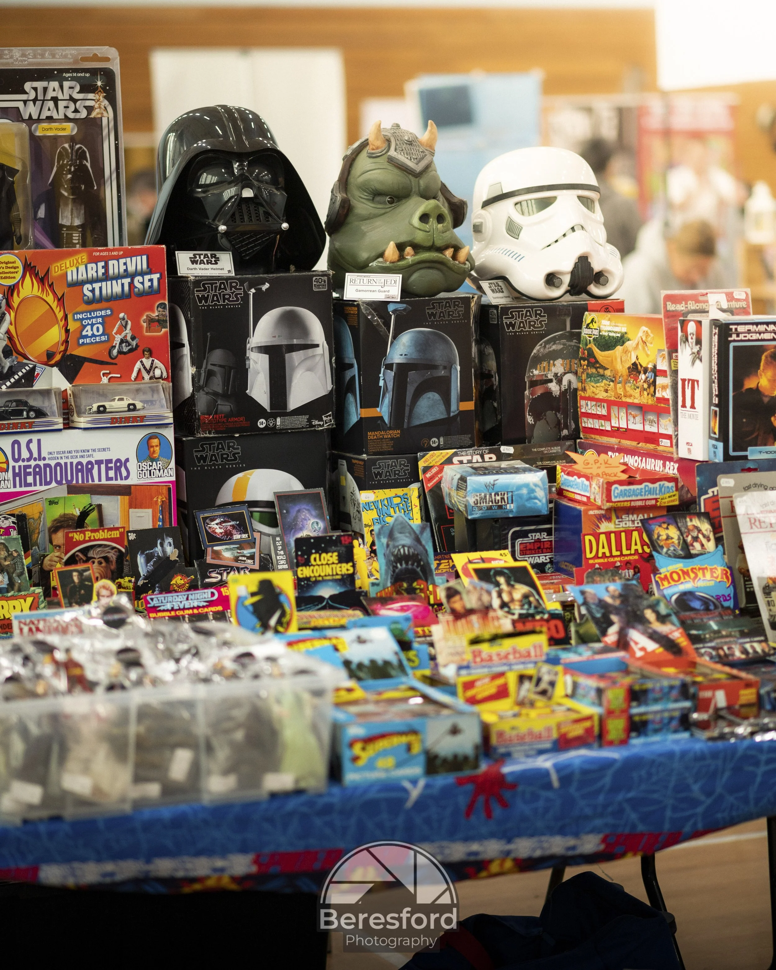 A table displays various collectible items and toys, including popular franchises like Star Wars and Jurassic Park, with helmets resembling Darth Vader, a Mandalorian helmet, and a Stormtrooper helmet, along with small boxes and packs of toys and games.