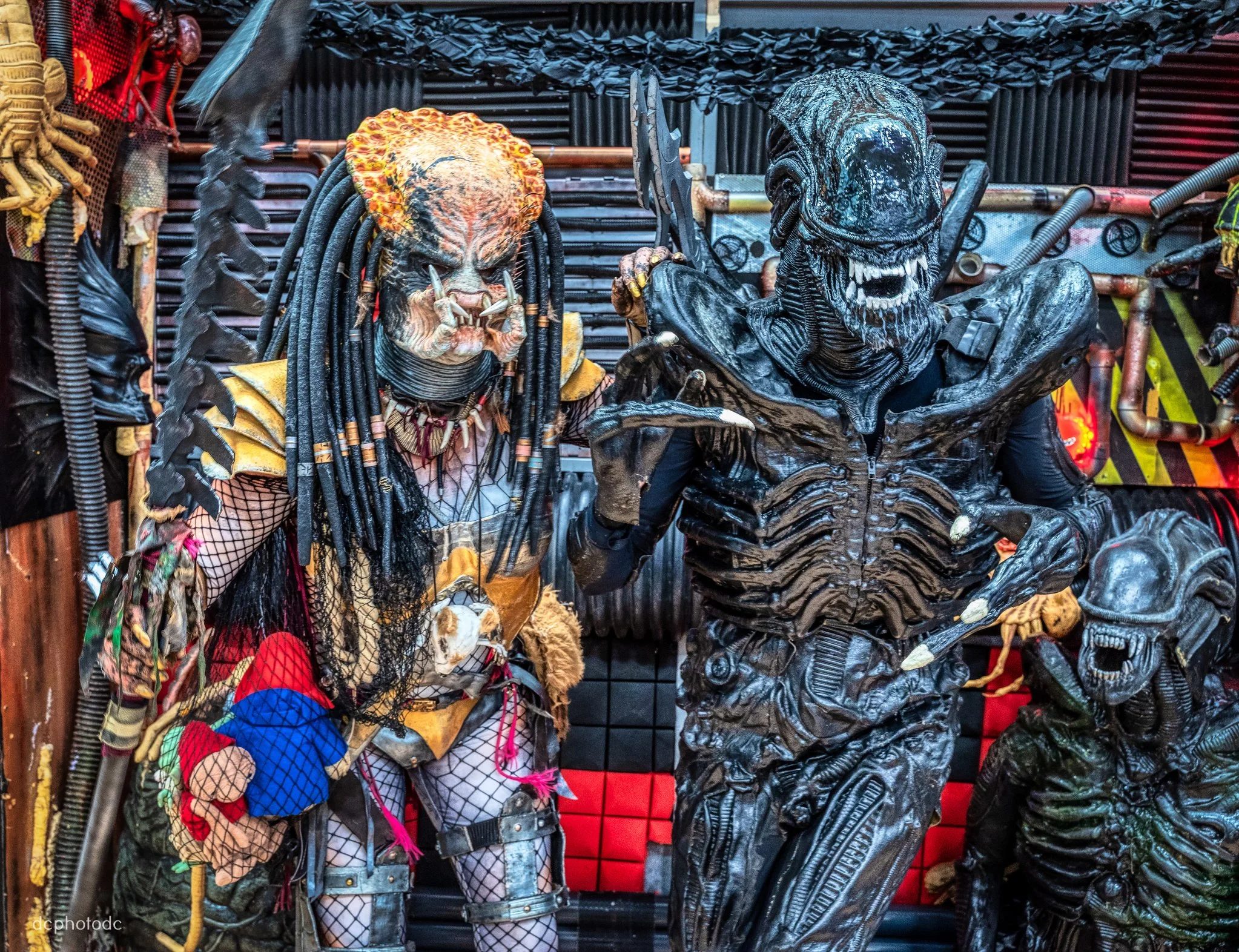 Two people dressed in detailed horror costumes resembling alien and predator characters, standing in front of a dark, industrial-themed background.