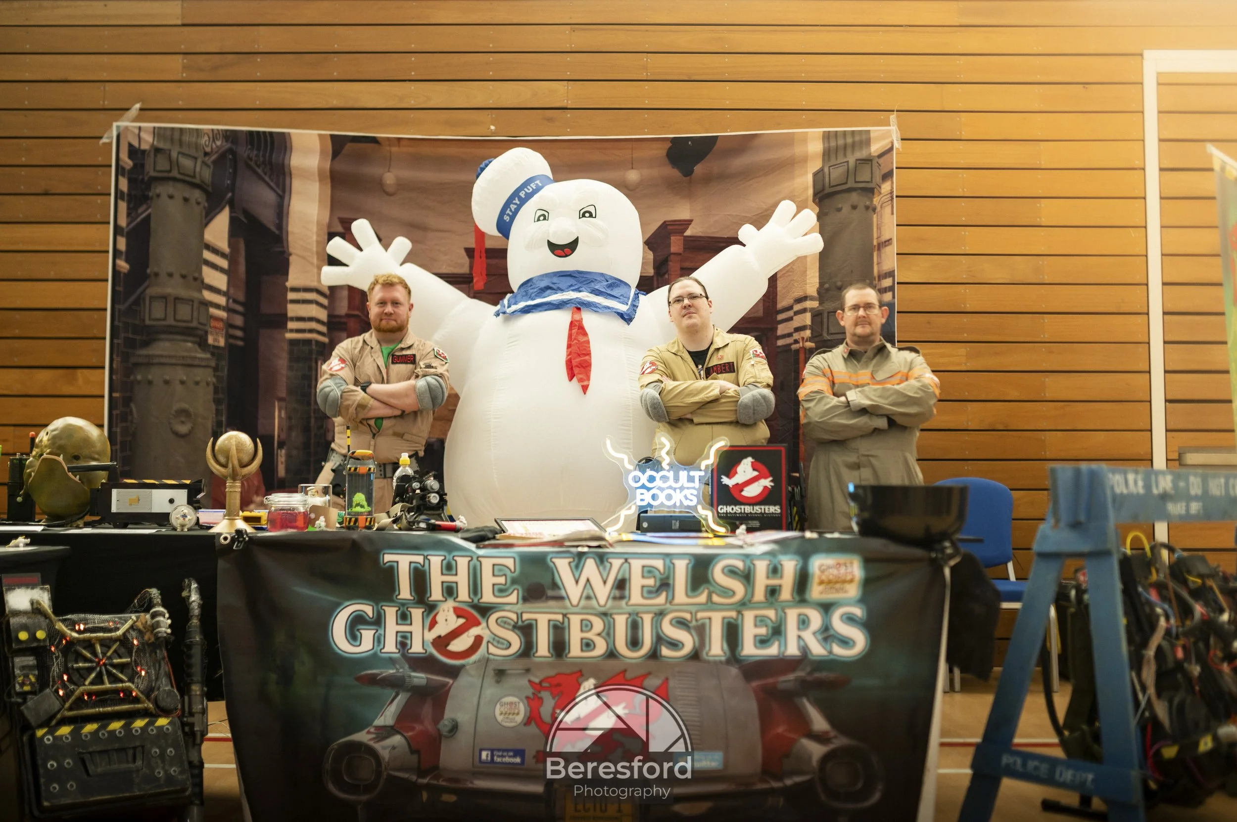 Three men standing with arms crossed behind a table displaying "The Welsh Ghostbusters" banner, next to a large inflatable marshmallow man from Ghostbusters, in a room with wooden walls.