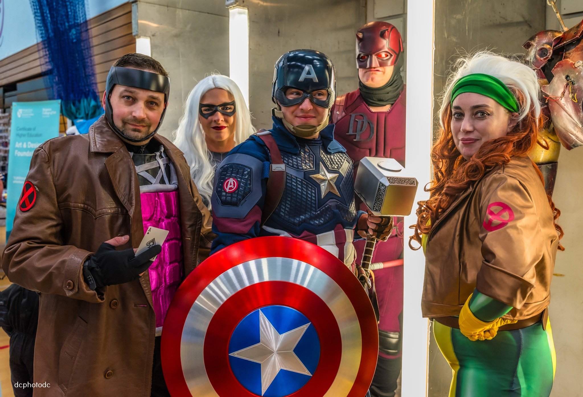Group of people in superhero costumes from Marvel, including Captain America and others, posing indoors with Marvel-themed props and artwork.