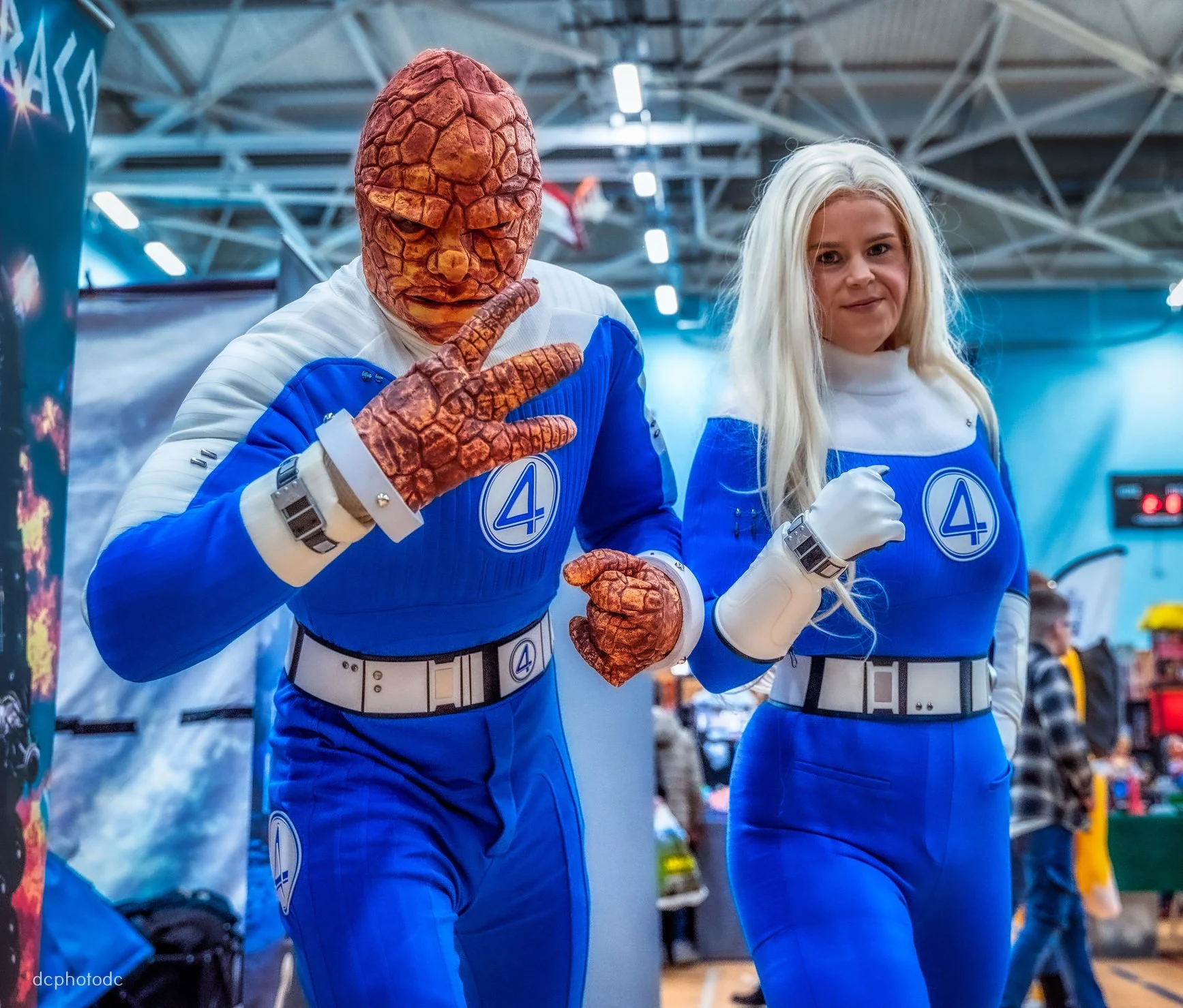 Two people in Fantastic Four costumes posing at a convention. The person on the left is wearing a Mr. Fantastic costume with a realistic orange mask, and the person on the right is wearing a Sue Storm costume with long blond hair. Both are in blue suits with the Fantastic Four logo.
