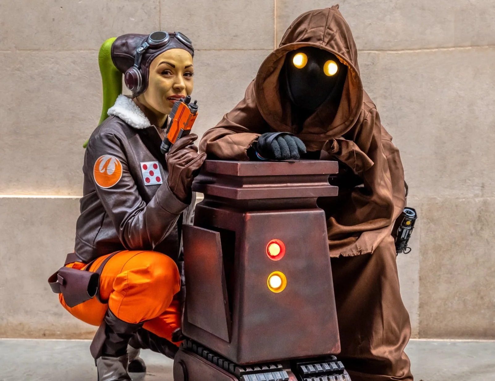 Two cosplayers dressed as characters from Star Wars, one in orange and brown gear holding a blaster, and the other in a brown hooded cloak with glowing yellow eyes, posing with a small robot.