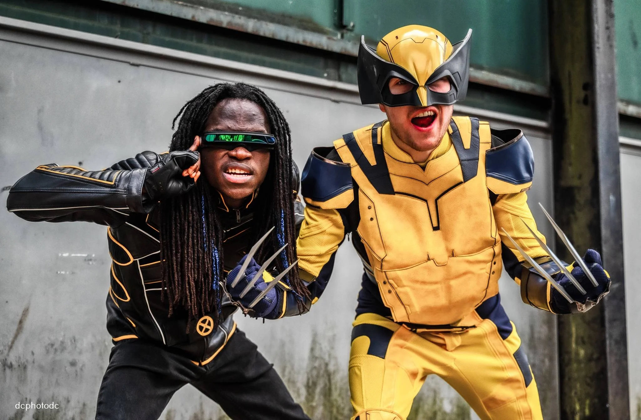 Two people dressed as superheroes posing with fierce expressions, wearing costumes and masks, one with long dreadlocks and the other with a yellow and black costume and mask, in an industrial setting.