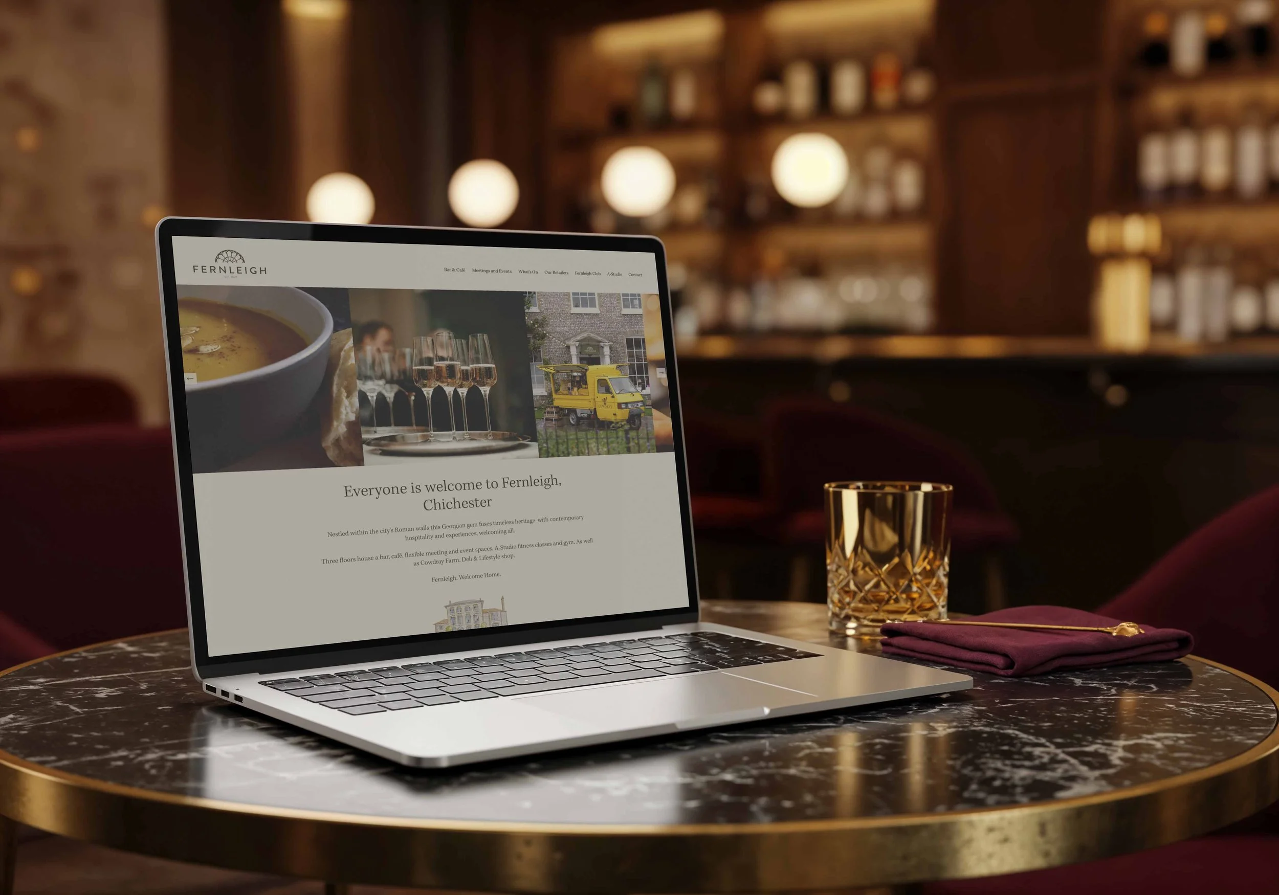 A laptop on a marble table displays a webpage for Fernleigh in Chichester, with images of a cup of soup, glassware, and a yellow van. A gold glass and a burgundy napkin with a gold stirrer are beside the laptop, set in a cozy, dimly lit bar or restau