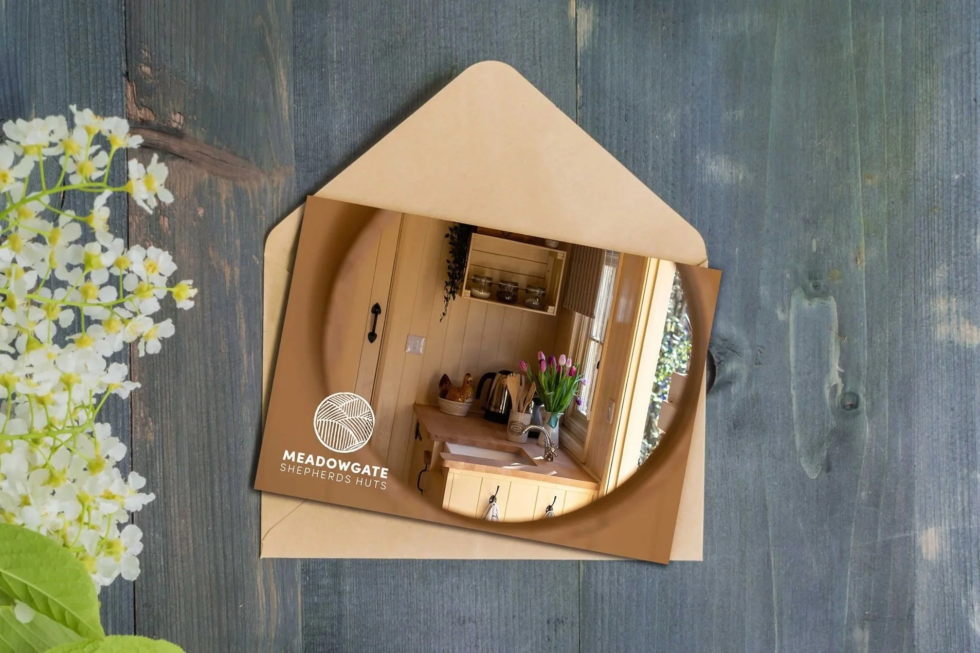 A mirror reflecting a cozy kitchen with wooden cabinets, a window with blinds, a vase of pink tulips, and kitchen appliances, on a rustic wooden surface next to white flowers and green leaves.