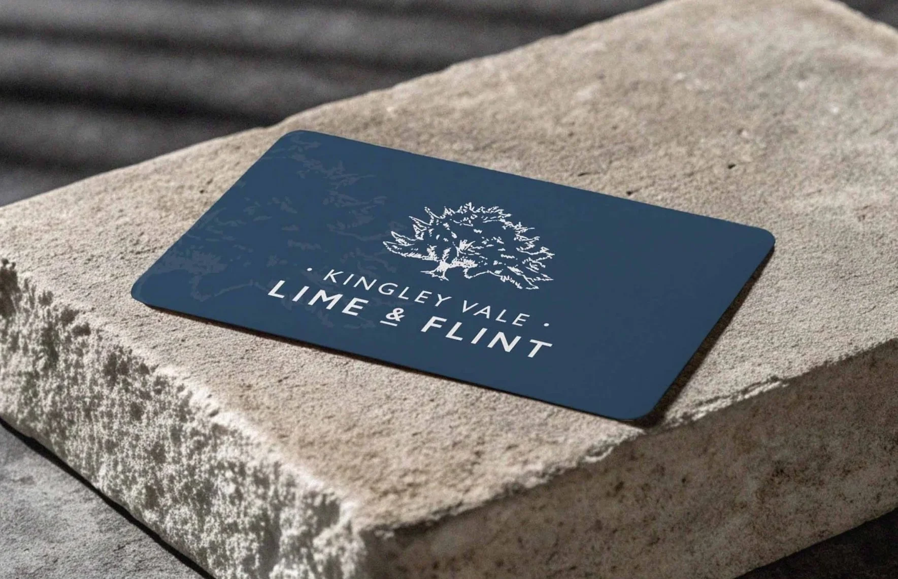 A blue business card on a light-colored stone surface. The card features a white tree graphic and the text 'KINGLEY VALE · LIME & FLINT'.