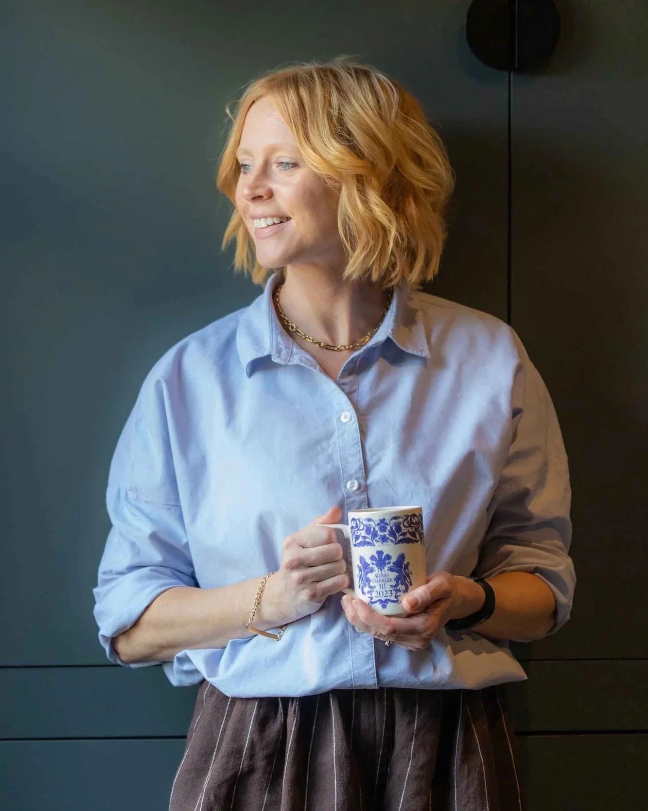A woman with short, blonde hair wearing a light blue button-up shirt and striped pants, holding a cup with a blue and white floral design and the year 2023 printed on it, smiling and looking to her left against a dark background.