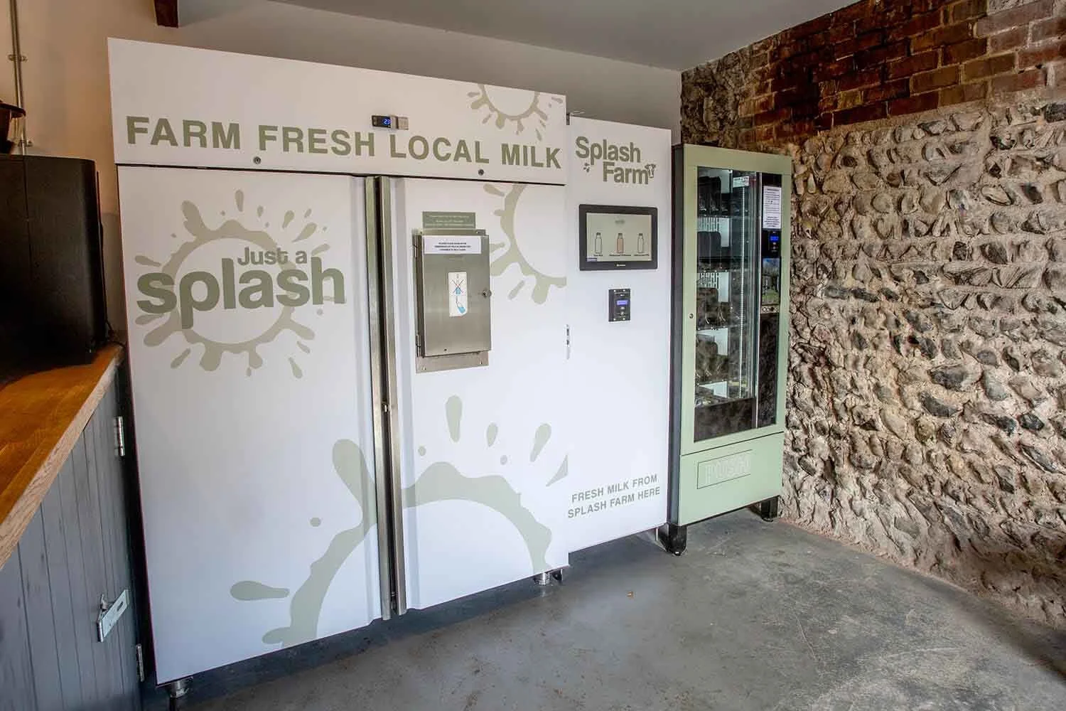 A milk vending machine labeled 'Splash Farm' offers farm fresh local milk. The machine features a white door with green text, a small locker, and a monitor showing milk options. There is also a green glass case containing bottled milk next to a stone