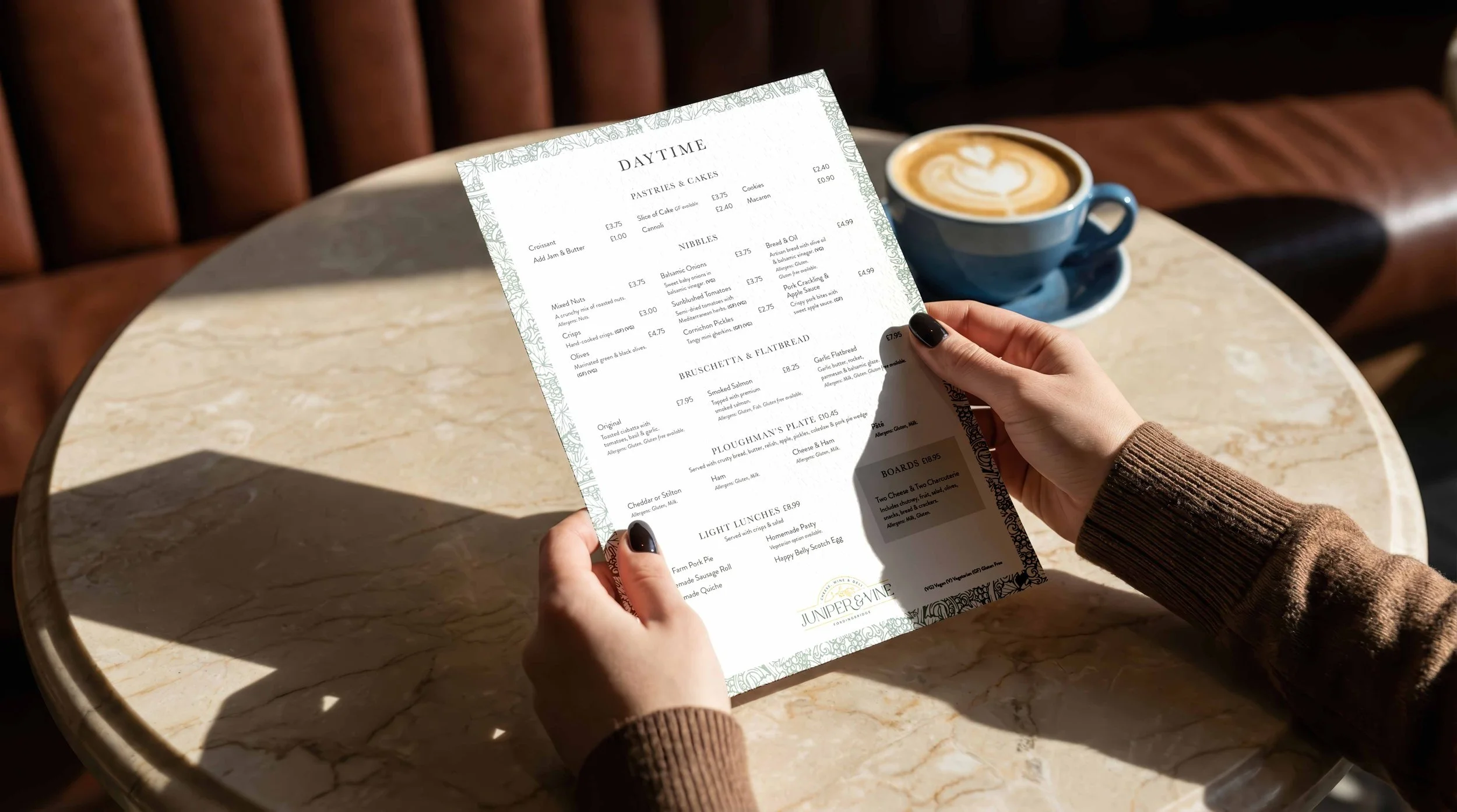 Person holding a printed cafe menu at a round table with a latte in a blue cup nearby, sunlight casting a shadow.