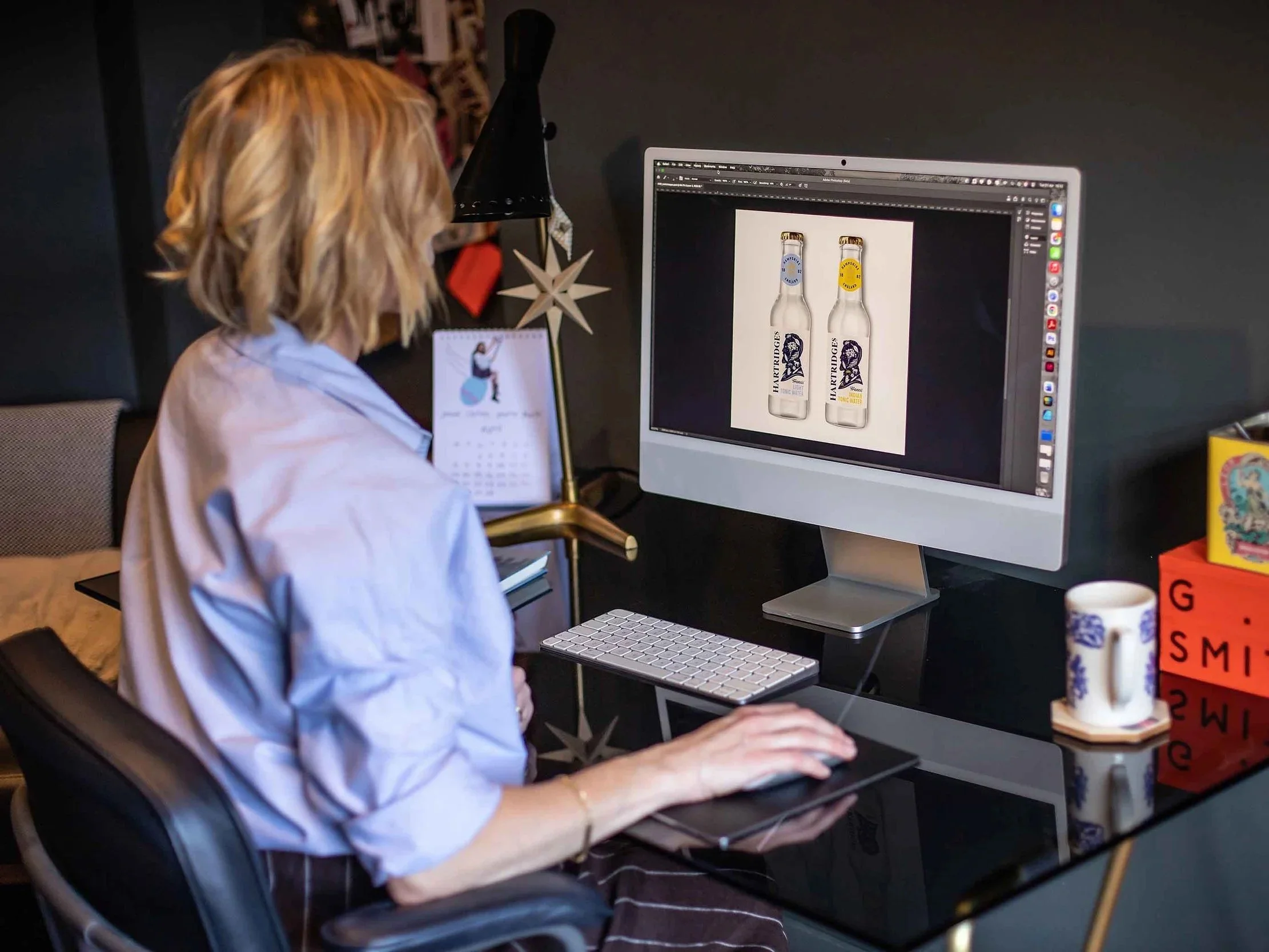 A woman with blonde hair working at a computer with a design of two glass bottles on the screen. She is sitting in a modern workspace, with various decorative items and a calendar nearby.