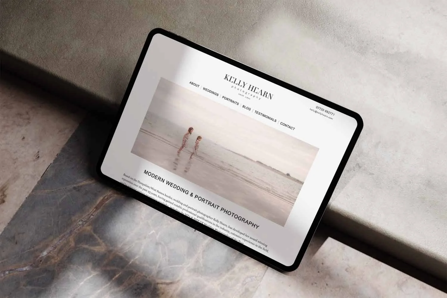 Tablet displaying a photography website named Kelly Hliban Photography, with menu options and a large photo of two children walking on a beach at sunset.