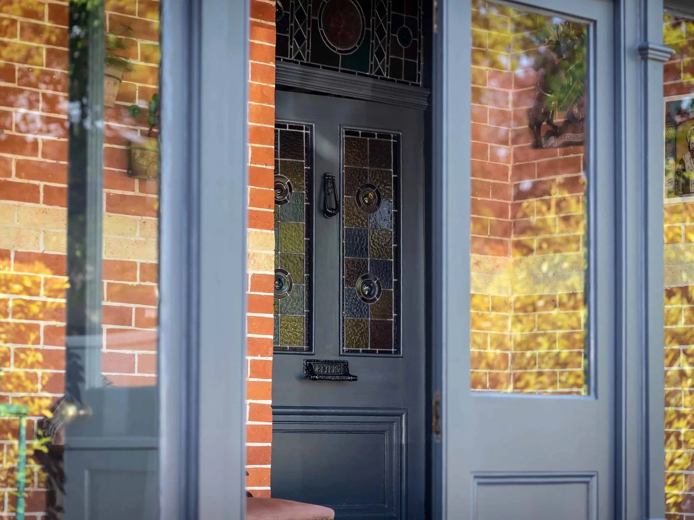 View through a window showing the front door of a house with a brick exterior, stained glass panels on the door, and a black handle. There are some plants and a decorative house plaque visible outside.