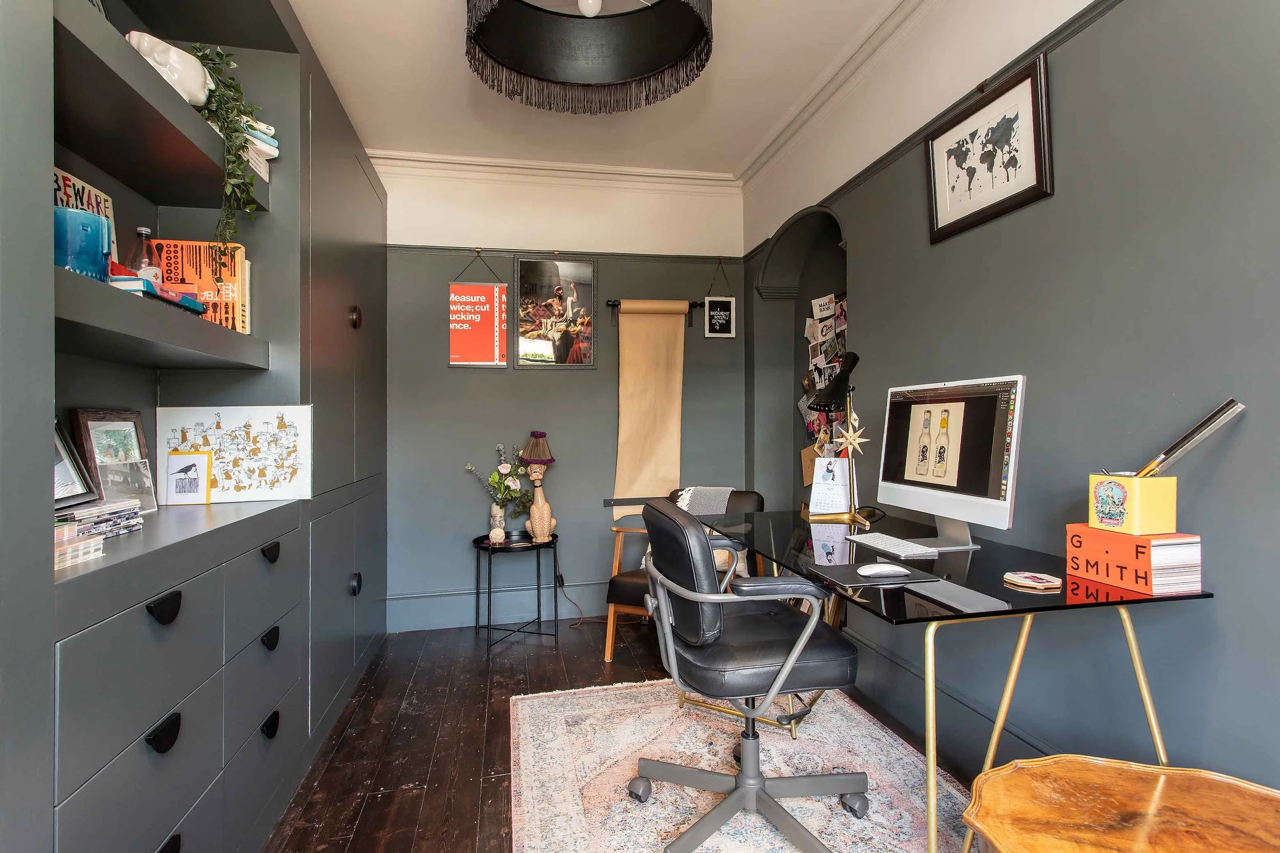 A home office with blue walls, a black desk with a computer, and various wall decorations including posters and artwork. There are chairs, a bookshelf with books and decorations, and a small apron hanger with photos and notes.