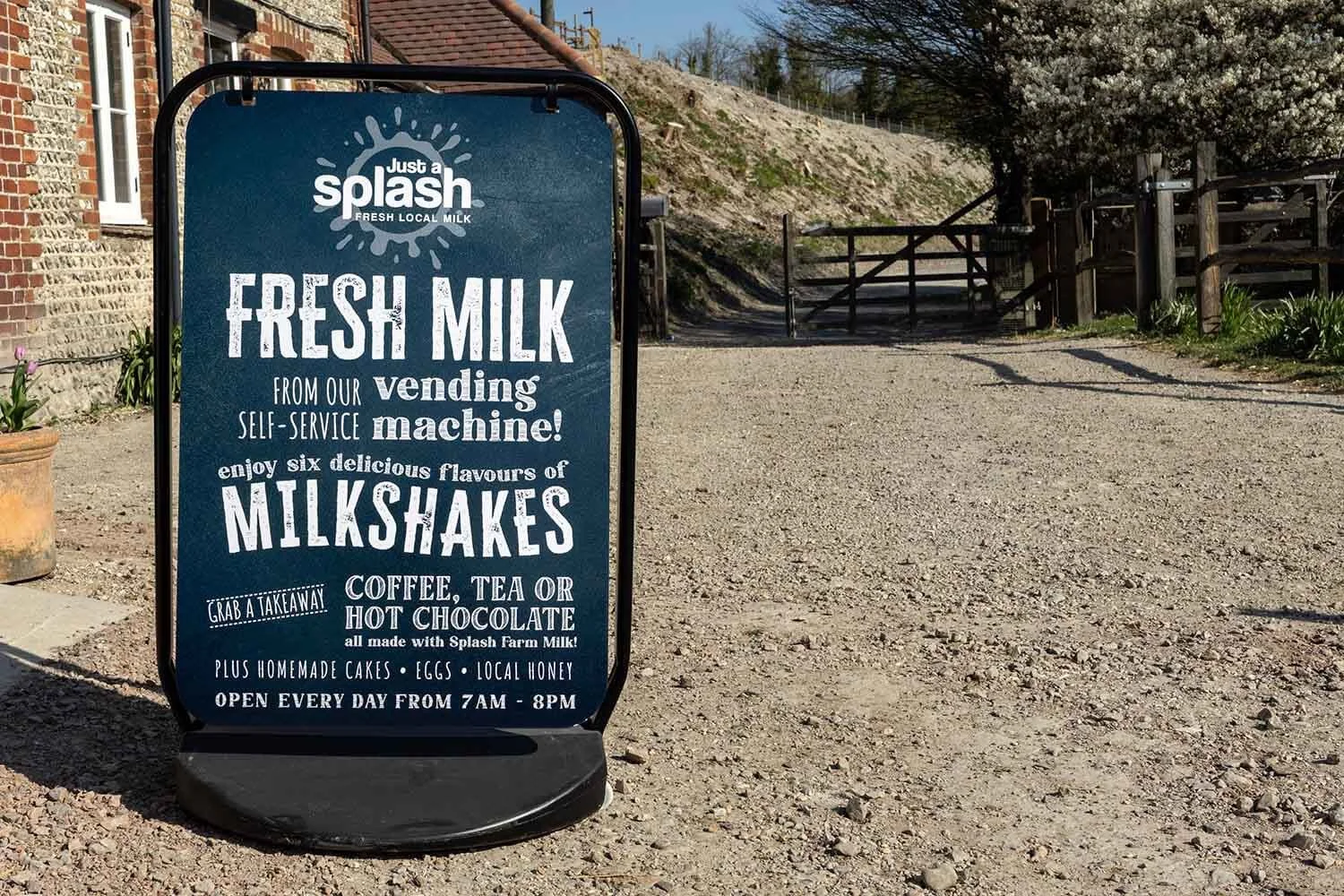 A blue outdoor signboard promoting fresh milk, milkshakes, and other homemade treats from Splash Farm Milk, available daily from 7 am to 8 pm, with a rustic outdoor setting including a dirt path, wooden fence, and blossoming trees in the background.