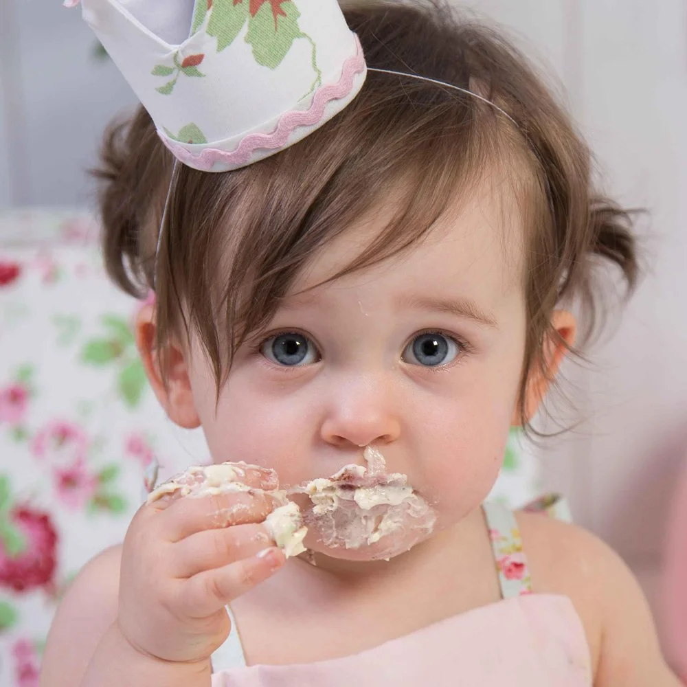 A young girl with blue eyes and brown hair, wearing a floral dress and a party hat, is eating a piece of cake.
