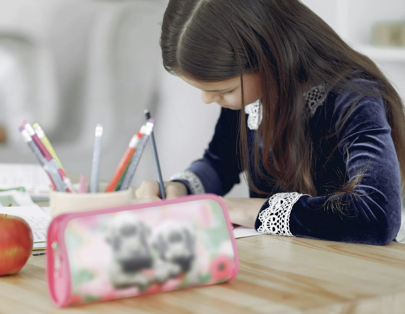 A young girl is studying at a desk with a pencil case in the foreground.