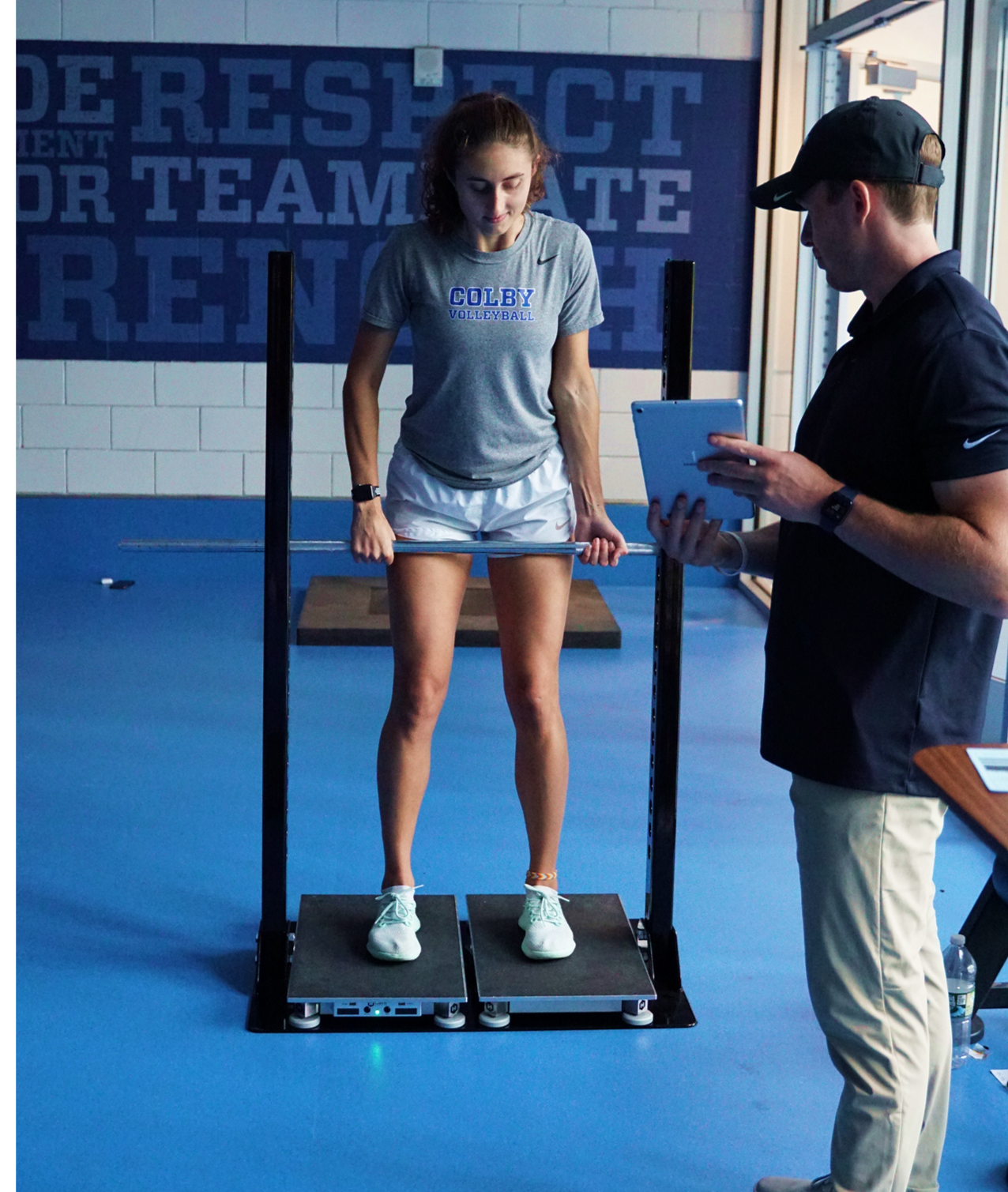 A female athlete standing on a force plate with a barbell, wearing a 'Colby Volleyball' shirt. A coach or instructor stands nearby holding a tablet. The room has motivational words on the wall.