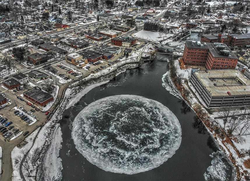 Aerial view of a cityscape with a circular ice formation in a river during winter, surrounded by buildings, roads, and snow-covered ground.