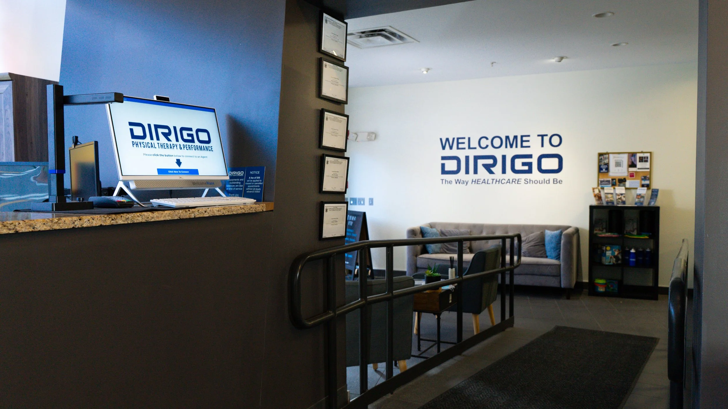 Reception area of a physical therapy clinic with a computer screen, framed certificates, and seating area.
