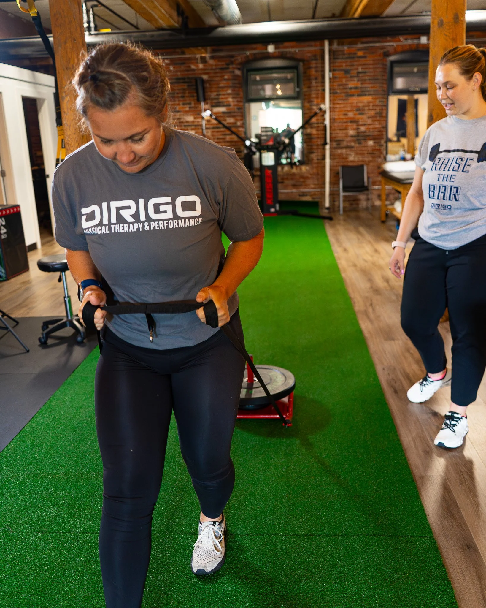 Woman draging a weighted sled on turf, getting physical therapy, a woman coaching her patient wearing a dirigo t shirt