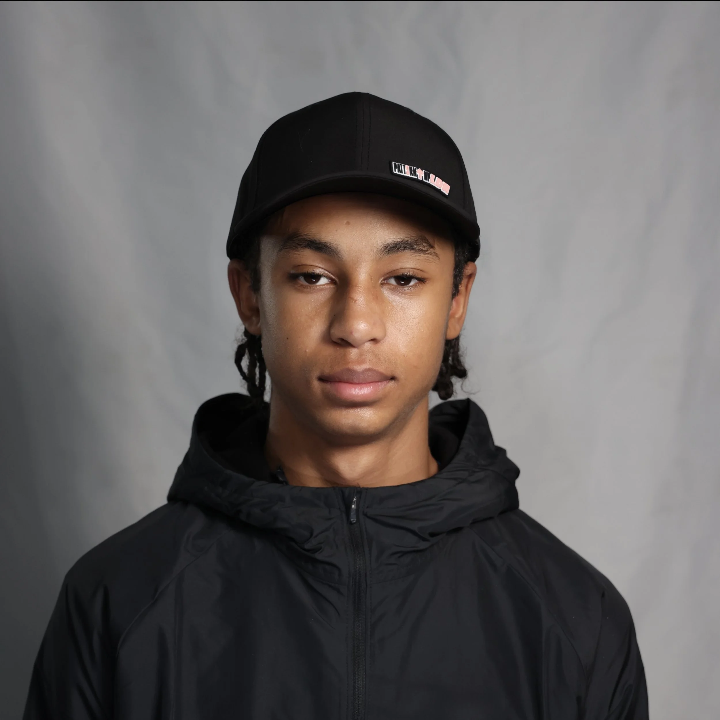 Portrait of a young man with curly hair wearing a black cap and black jacket, looking directly at the camera against a neutral background.